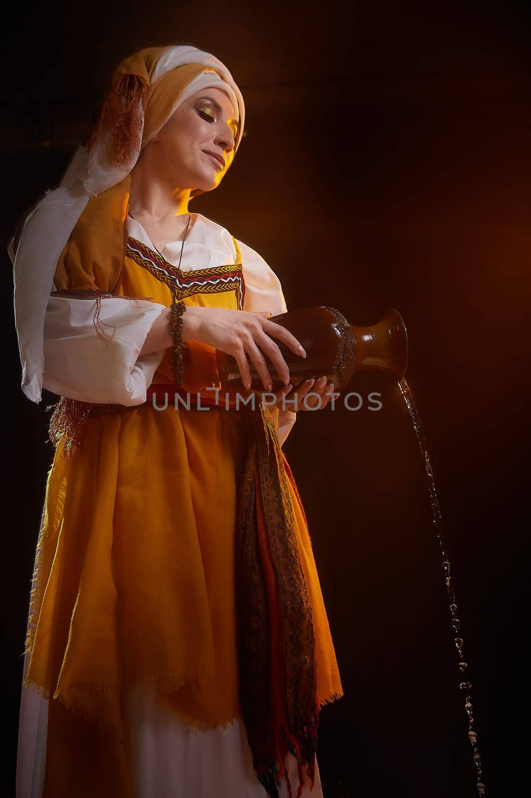 A girl in traditional eastern attire gracefully pours water from a clay pot, illuminated by warm, dramatic lighting.