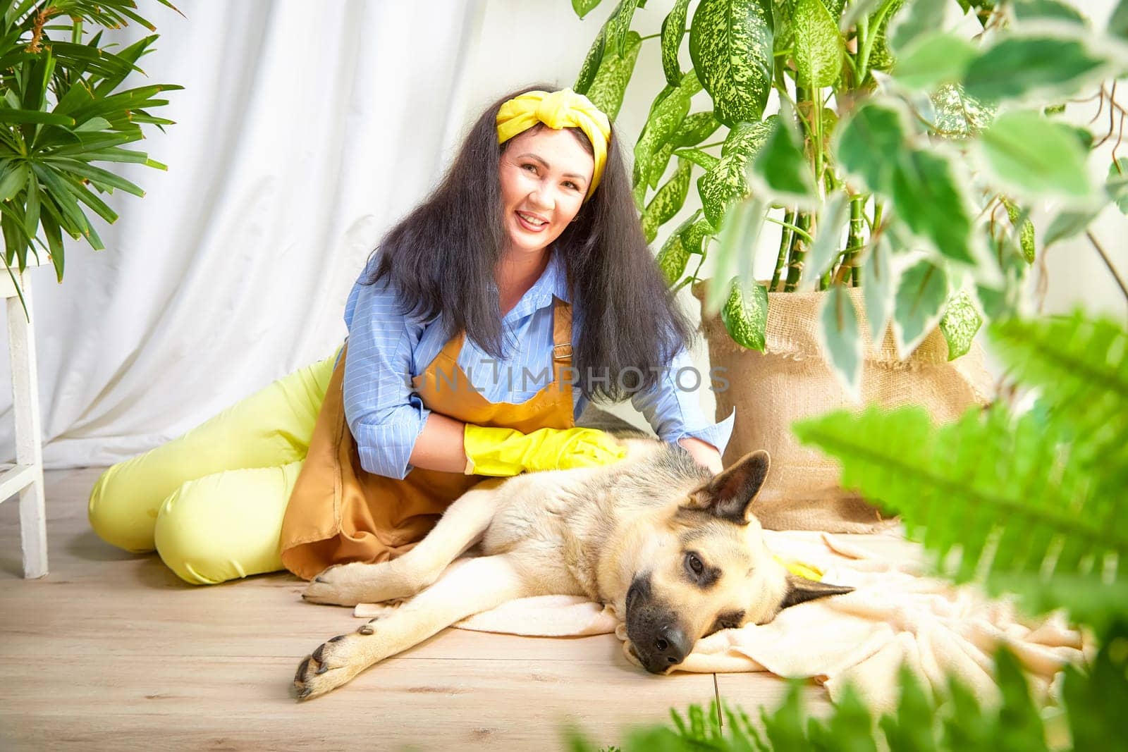 A gardener smiles while caring for flowers alongside a relaxed dog in a greenhouse.