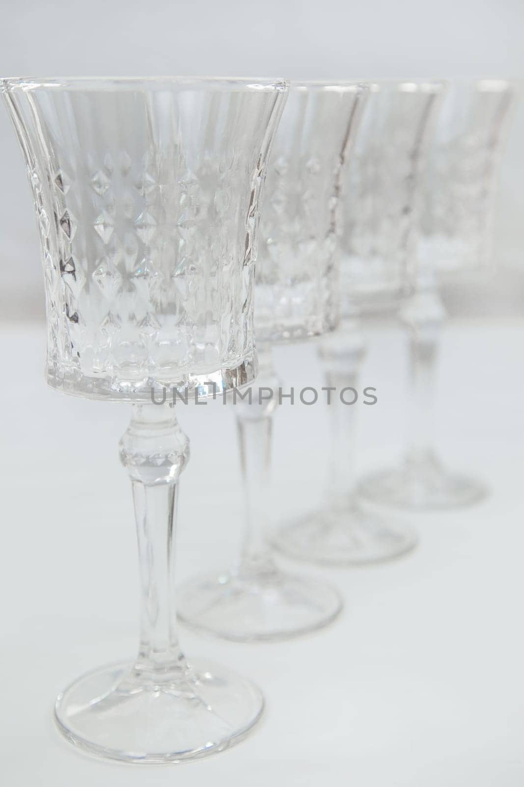 Crystal wine glasses on a long stem, with expanded edges and a faceted pattern stand in a row on a white background, the first one is in focus on the sides, the second ones stand behind it in focus. High quality photo