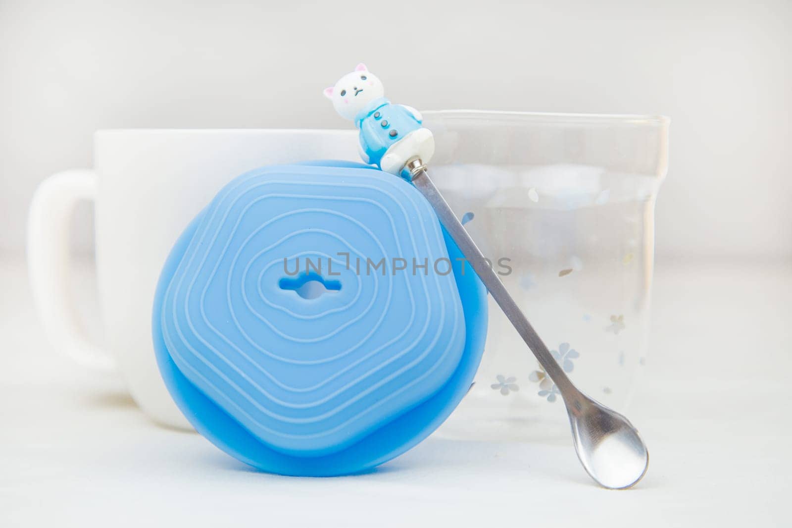 A white mug with a teaspoon with a figure on the handle of a spoon in the form of a rabbit, a blue lid and a transparent glass stands on a white background. High quality photo