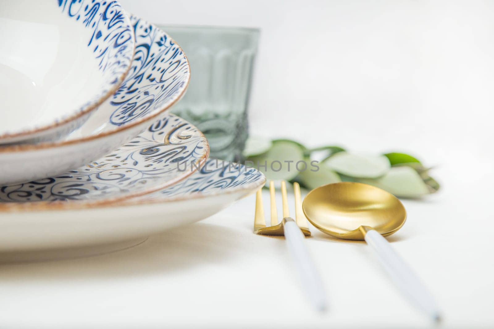 On the side there is a set of plates of different sizes with a white middle and blue patterns on the side of the plate and a gold border, they stand on top of each other with a tilt, there are gold - white devices on the side, there is a glass and green branches of plants, a side view of a close-up on a white background. The concept of serving. The concept of eating. High quality photo