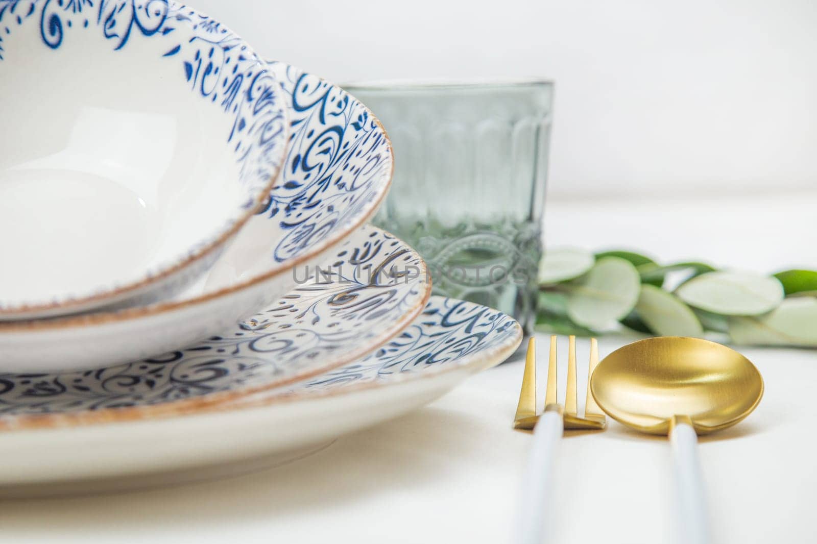 On the side there is a set of plates of different sizes with a white middle and blue patterns on the side of the plate and a gold border, they stand on top of each other with a tilt, there are gold - white devices on the side, there is a glass and green branches of plants, a side view of a close-up on a white background. The concept of serving. The concept of eating. High quality photo
