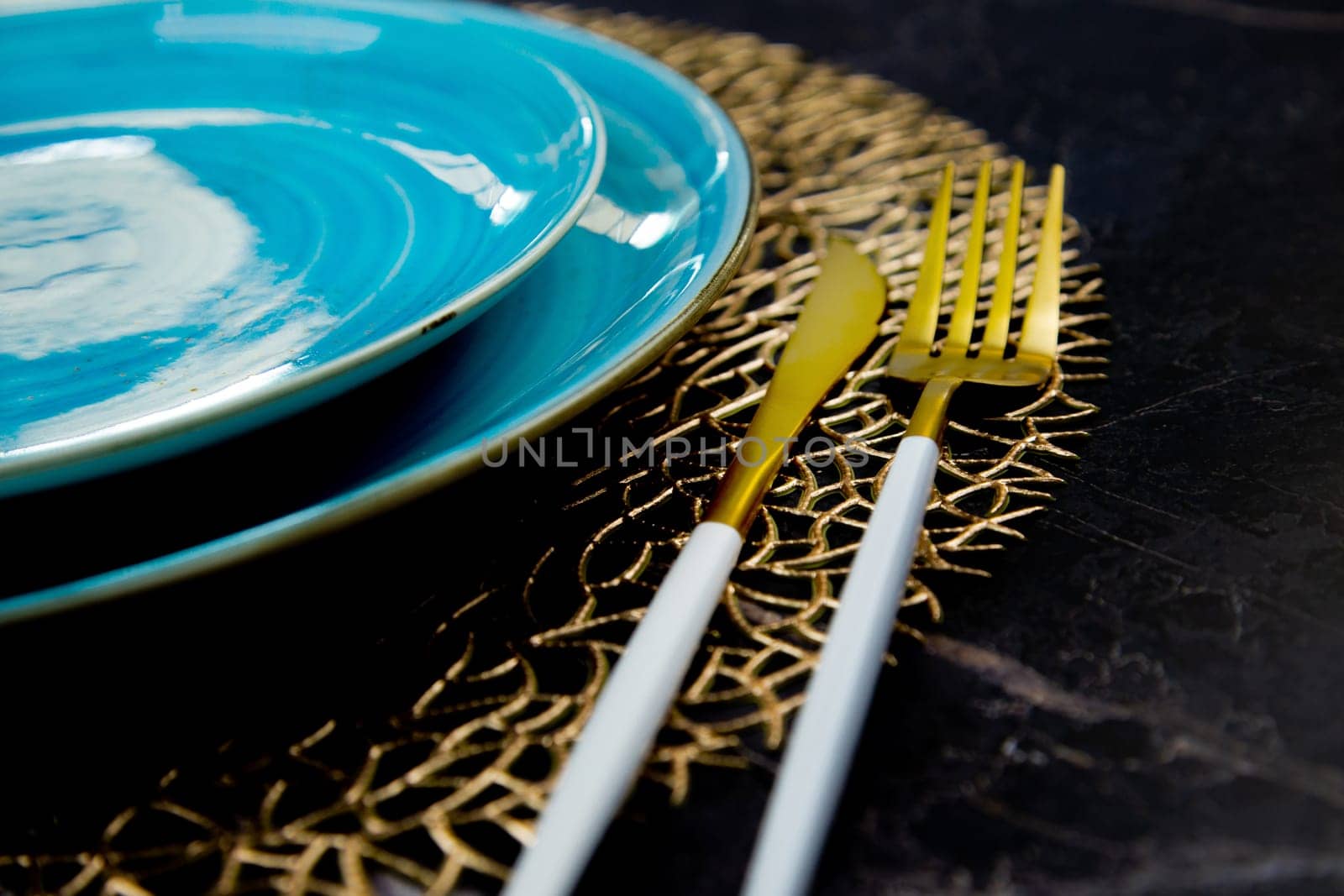 Close-up. View of the part of the blue plates stand one on one on a gold patterned napkin, gold cutlery lies on the side: a fork and a knife on a black background. The concept of serving, the culture of eating. High quality photo