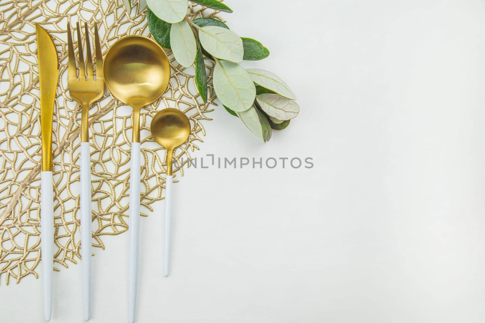 Cutlery: a knife, fork, spoon and a teaspoon of gold color with a white handle lie on a napkin of gold color, next to a green twig on a white background. The concept of food, culture and eating. . High quality photo