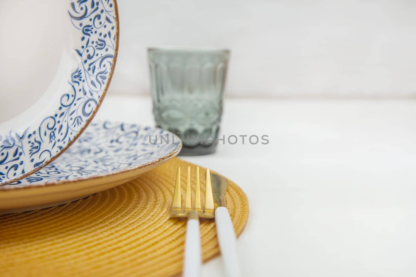 A white soup plate with a blue pattern on the edges stands directly on a blue-and-white patterned serving plate in close-up on a yellow straw napkin, a knife and fork are cutlery next to them, a blue glass on a white background is visible behind them. The concept of eating, table setting. High quality photo