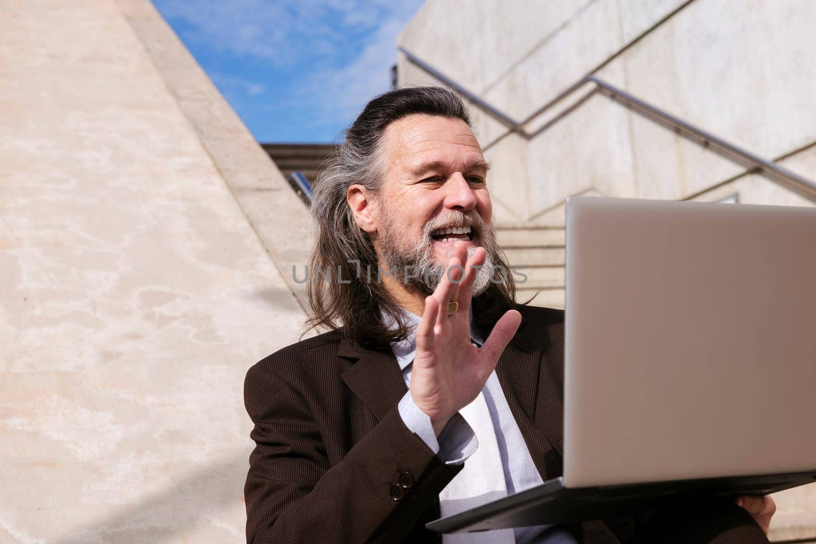 positive middle age male entrepreneur in elegant suit siting on stairway and waving hand saying hello while having online video meeting via laptop, concept of business and middle age man lifestyle