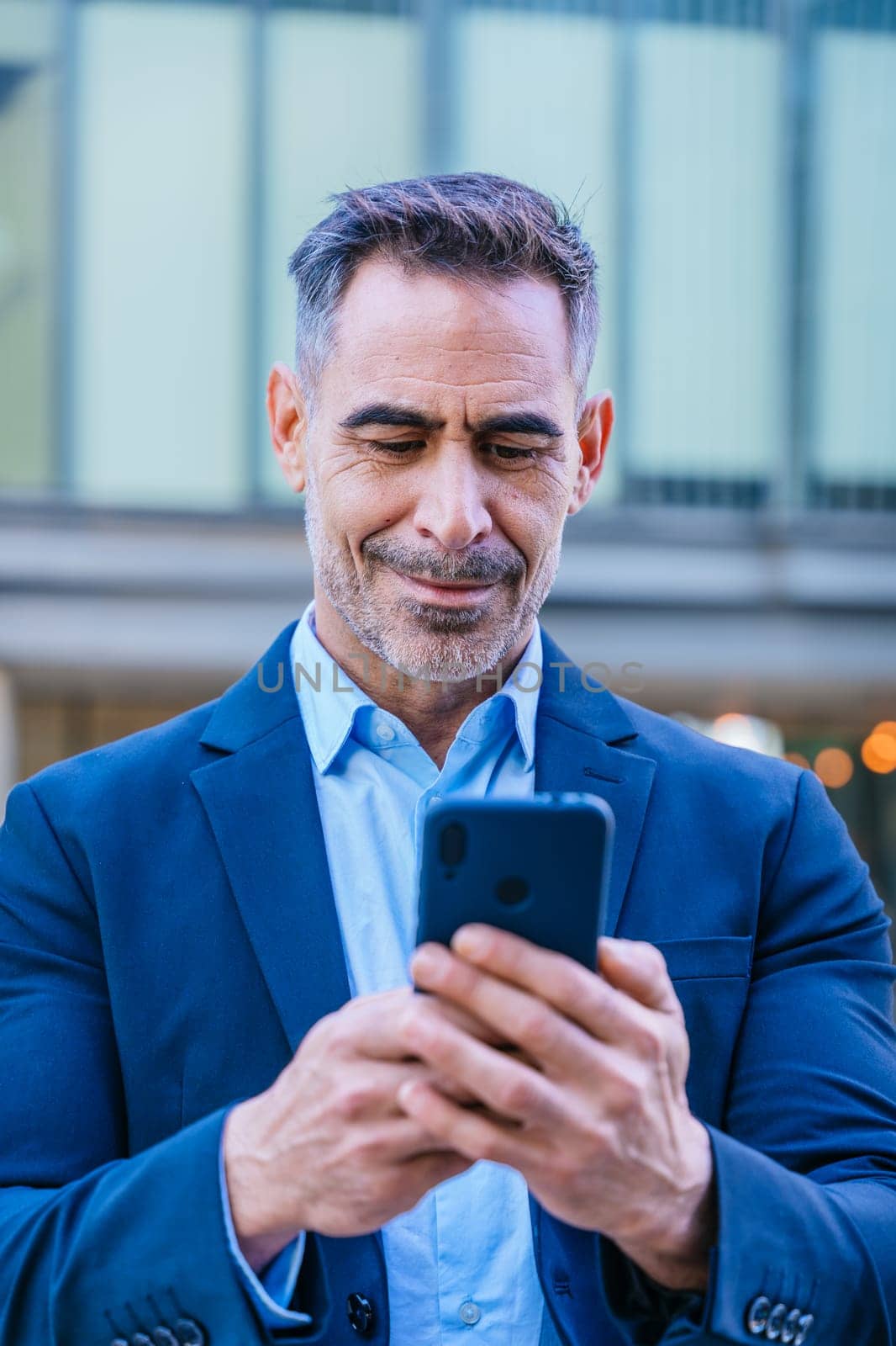 Smiling middle-aged businessman in a blue suit holding and using a smartphone outdoors in a modern urban setting.