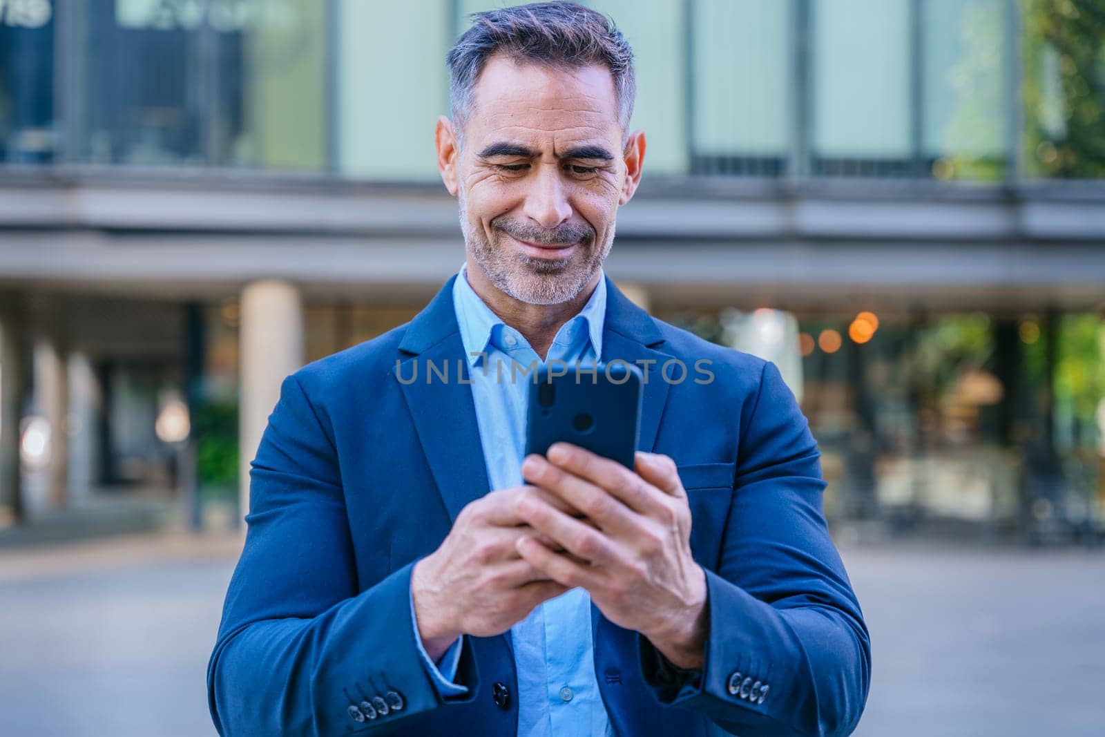 Smiling middle-aged businessman in a blue suit holding and using a smartphone outdoors in a modern urban setting.
