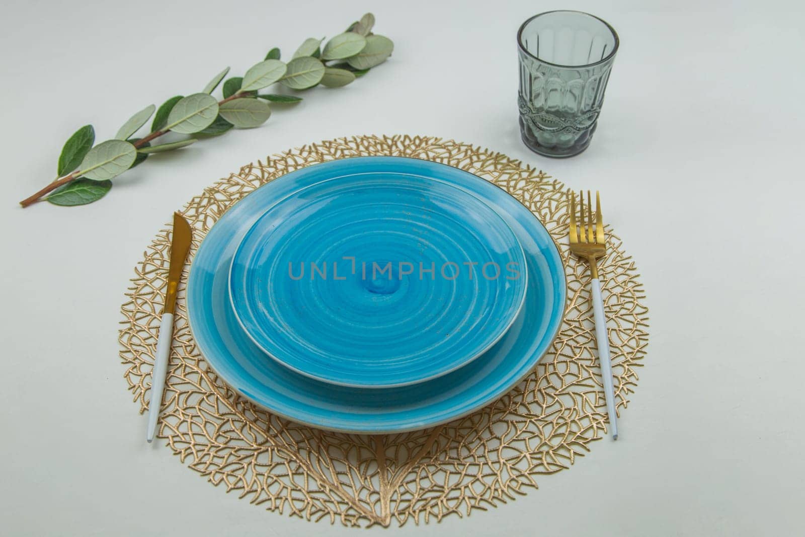 Two bright blue serving plates with a spiral pattern of different sizes, stand one on one on a golden textured napkin, serving devices of gold - white color are located on the side, there is a glass on top and a green branch of the plant lies sideways, side view on a white background. The concept of serving. The concept of eating. High quality photo