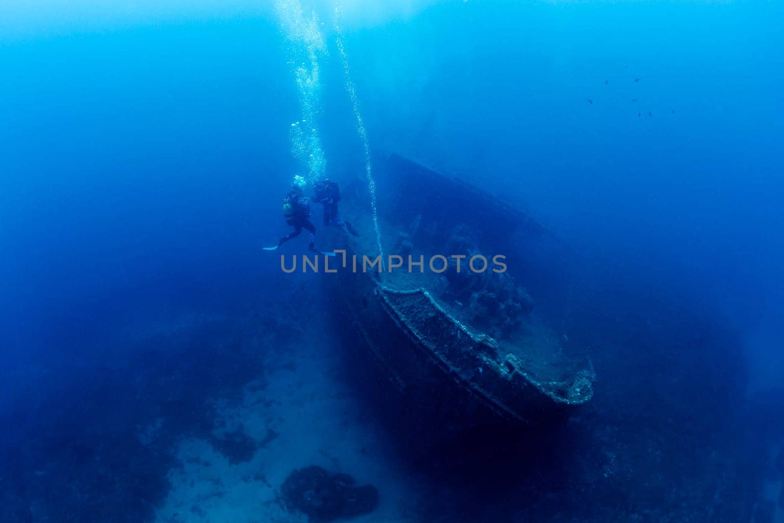 two scuba divers diving near of the wreck of an old ruined ship on the bottom of deep blue sea with clear water and light glare, concept of underwater adventure and seascape