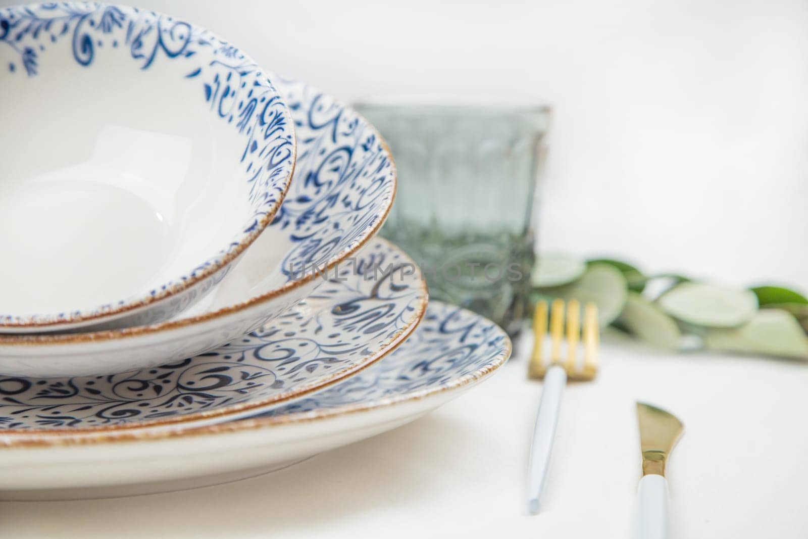 On the side there is a set of plates of different sizes with a white middle and blue patterns on the side of the plate and a gold border, they stand on top of each other with a tilt, there are gold - white devices on the side, there is a glass and green branches of plants, a side view of a close-up on a white background. The concept of serving. The concept of eating. High quality photo