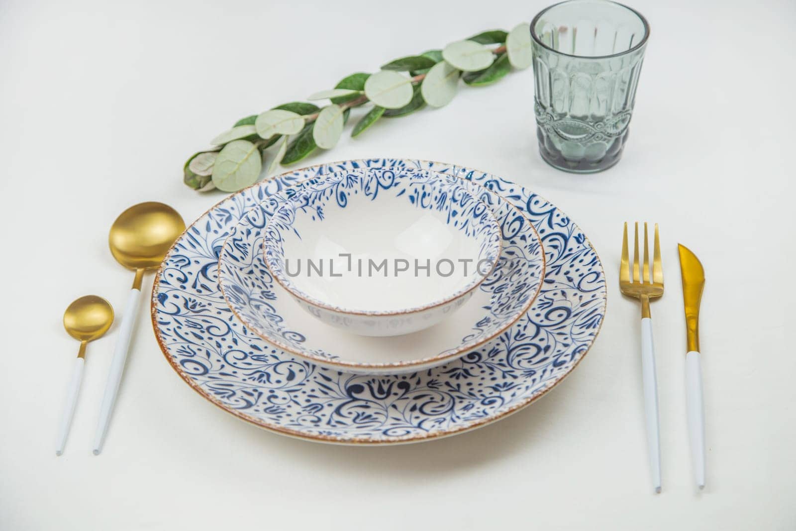 A set of white plates with a blue pattern on the edges stand on top of each other, spoons, a knife and a fork lie on the sides, behind them is a blue glass and a green sprig of sprouting on a white background. The concept of eating, table setting. High quality photo