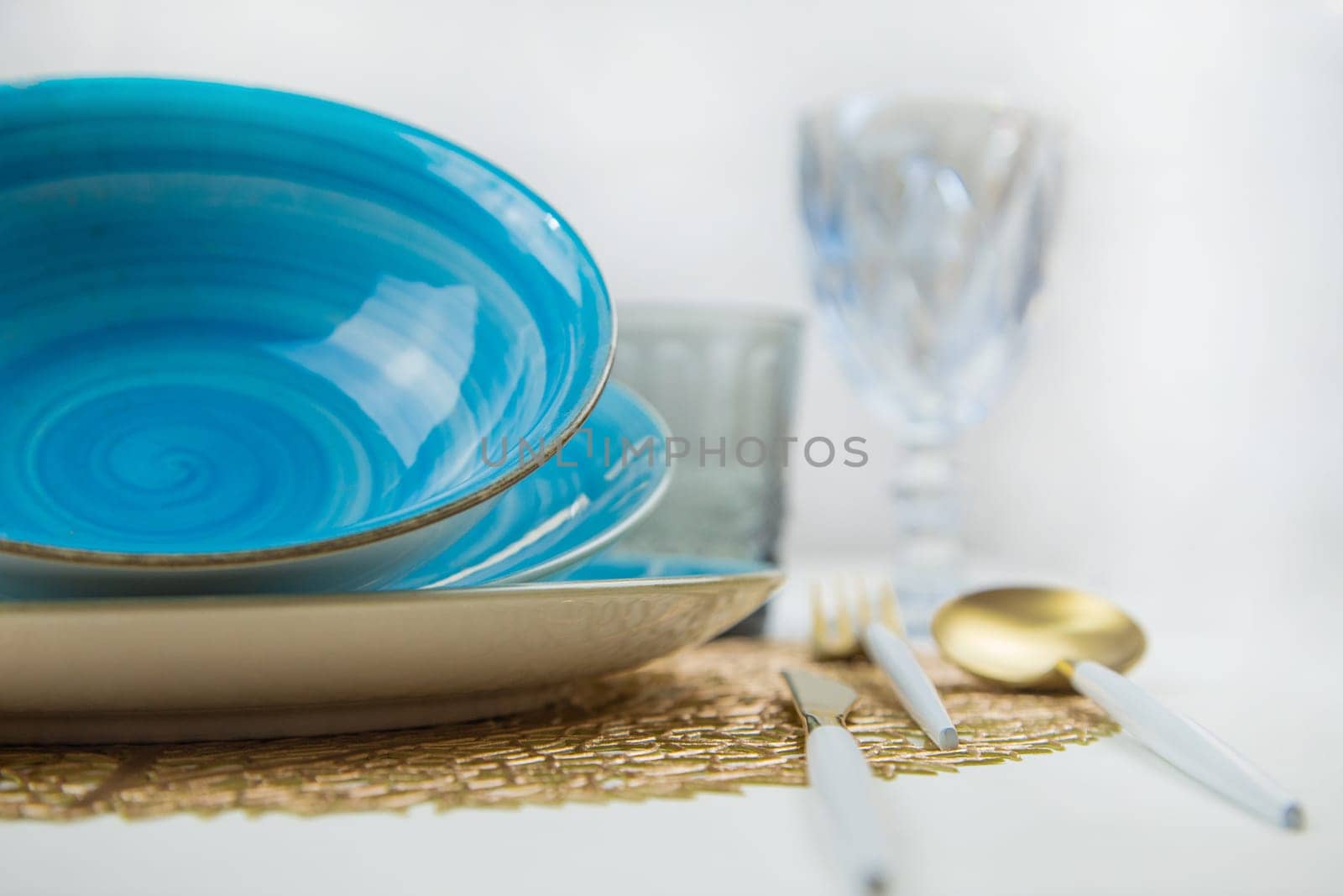 Bright blue serving plates with a spiral pattern, tilted one after the other, serving devices of gold - white color are located on the side, glasses and glasses are blurred behind them, the side view is large on a white background. The concept of serving. The concept of eating. High quality photo