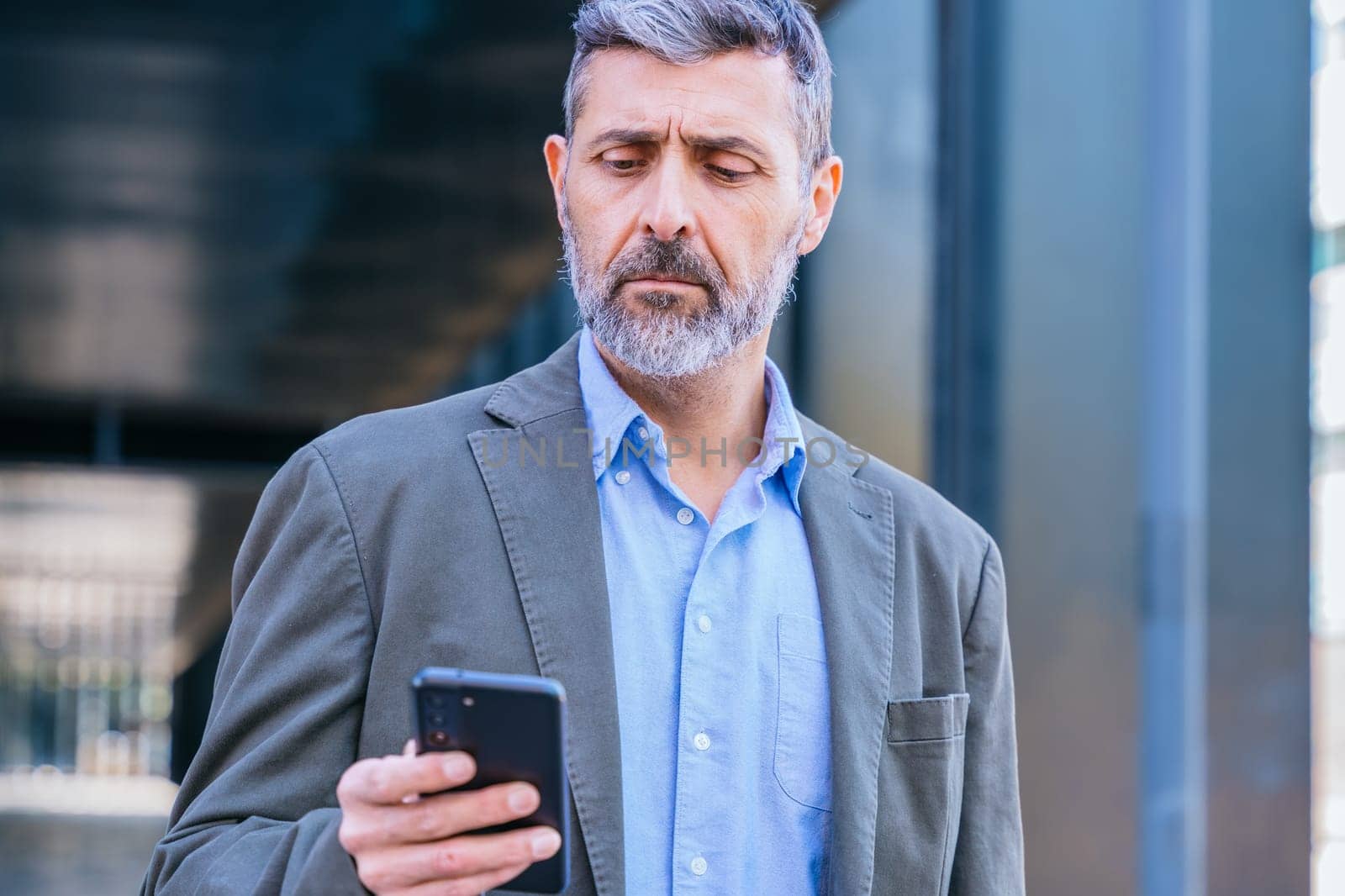 Smiling middle-aged businessman in a blue shirt holding and using a smartphone outdoors in a modern urban setting.