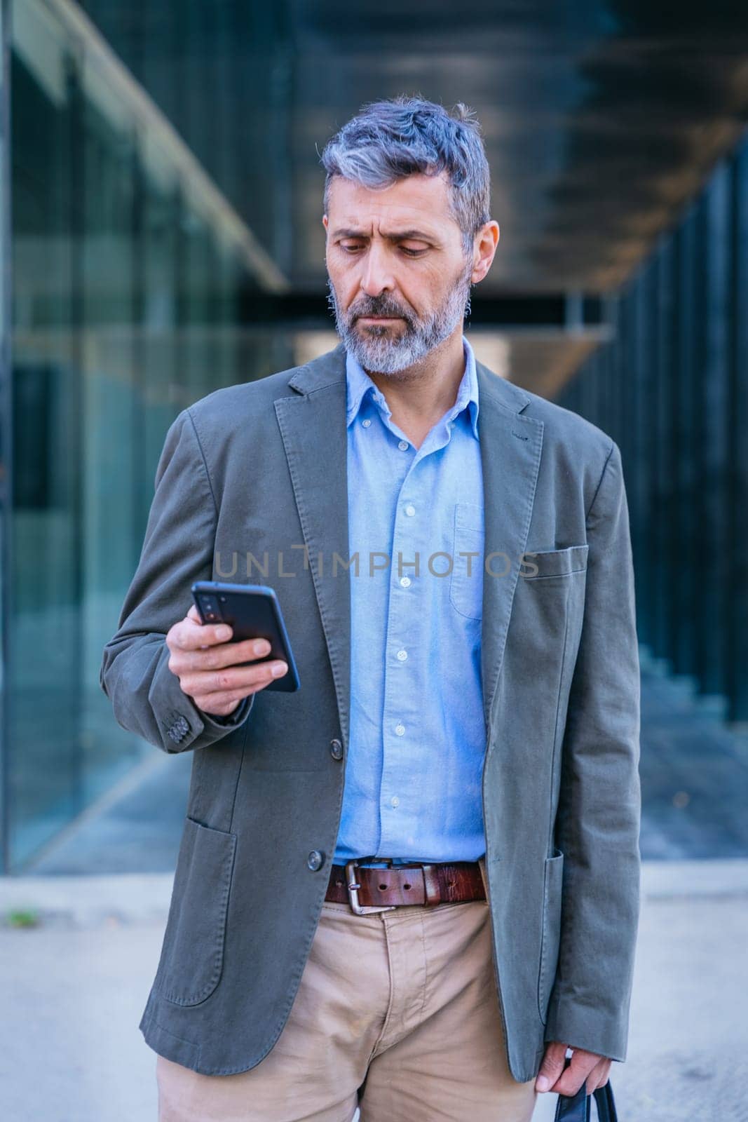 Smiling middle-aged businessman in a blue shirt holding and using a smartphone outdoors in a modern urban setting.
