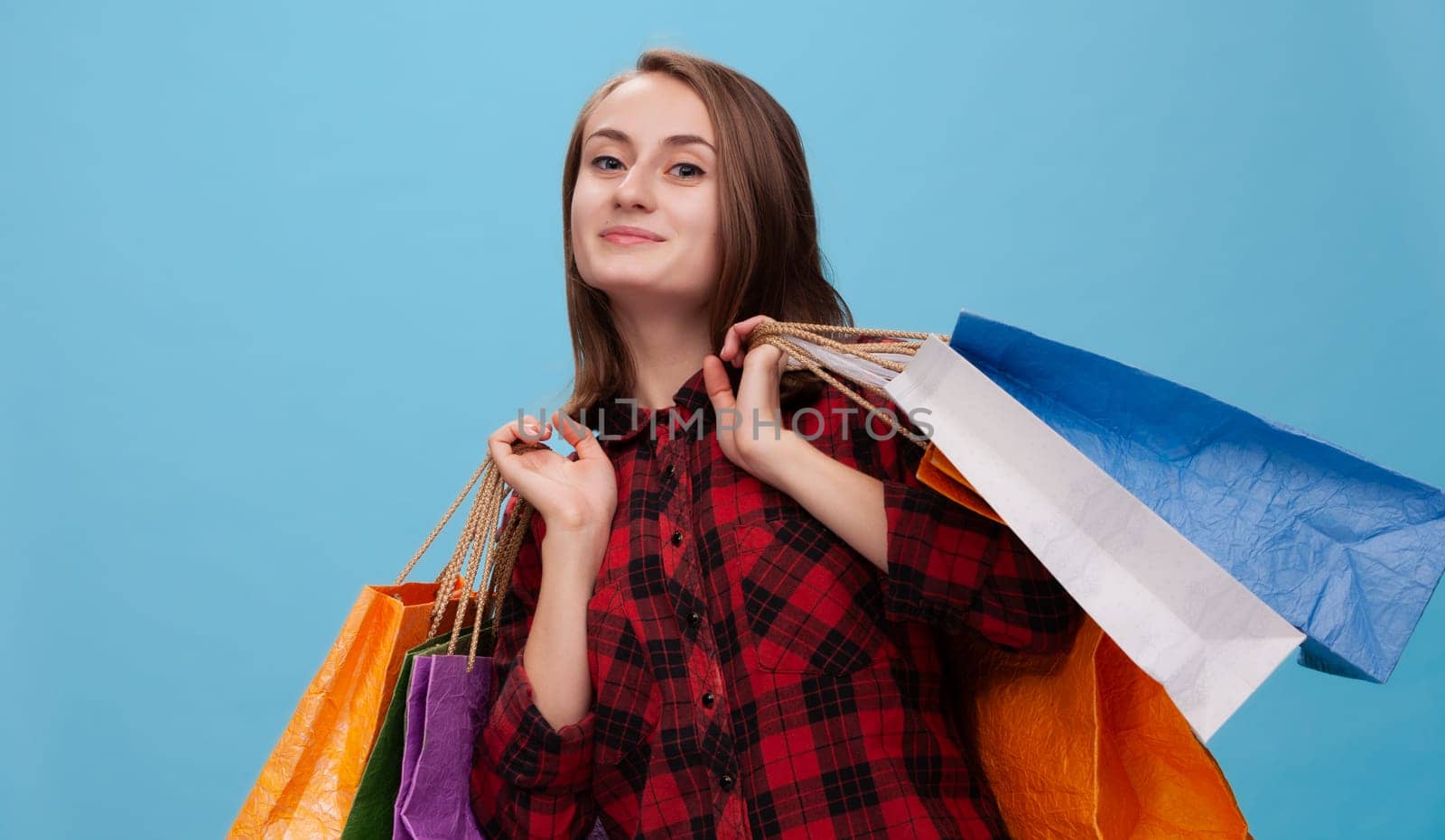 A joyful young woman stands against a blue background, holding multiple shopping bags in vibrant colors. She radiates positivity, showcasing the excitement of shopping during the commerce season.