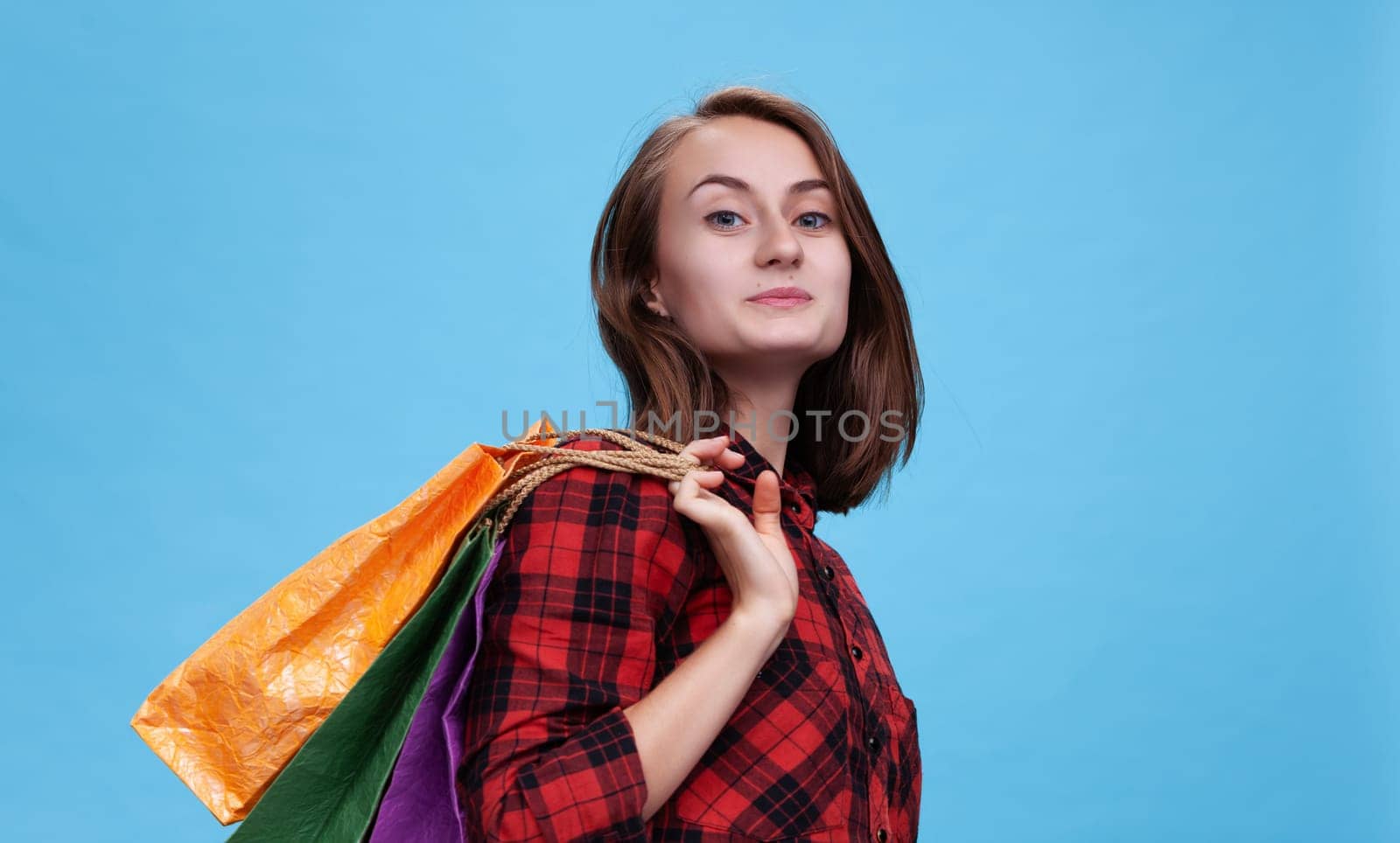 A cheerful young woman stands confidently against a blue backdrop, showcasing her vibrant shopping bags. She embodies the joy of the commerce season, ready for an exciting day.