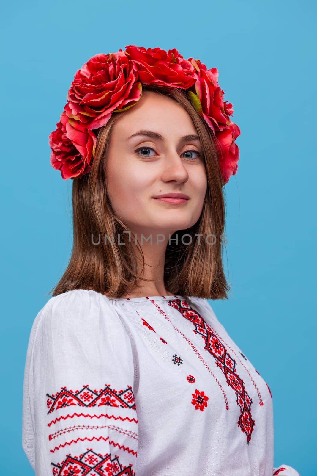 A girl stands confidently against a blue backdrop, showcasing her traditional Ukrainian embroidered shirt. Her floral headpiece adds a touch of color and culture to her appearance.