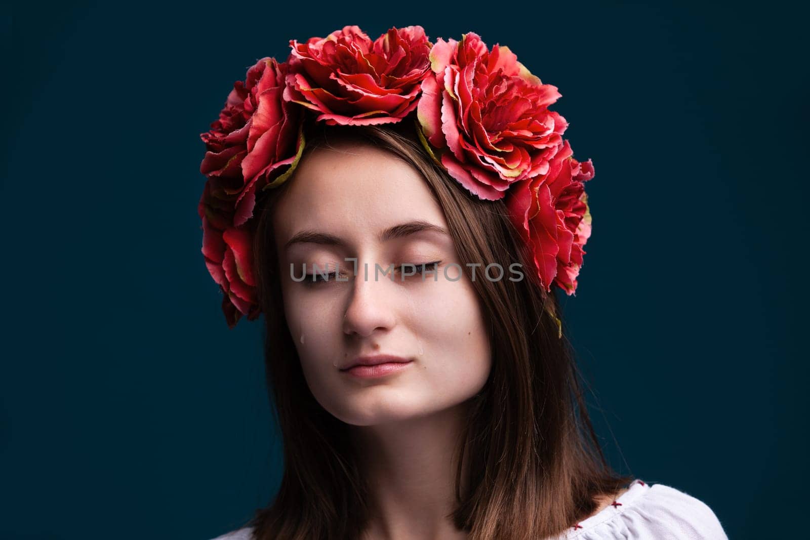 A young woman stands with her eyes closed, tears on her cheeks. She wears a crown of vibrant red flowers, surrounded by a dark background, creating a poignant and emotional atmosphere.