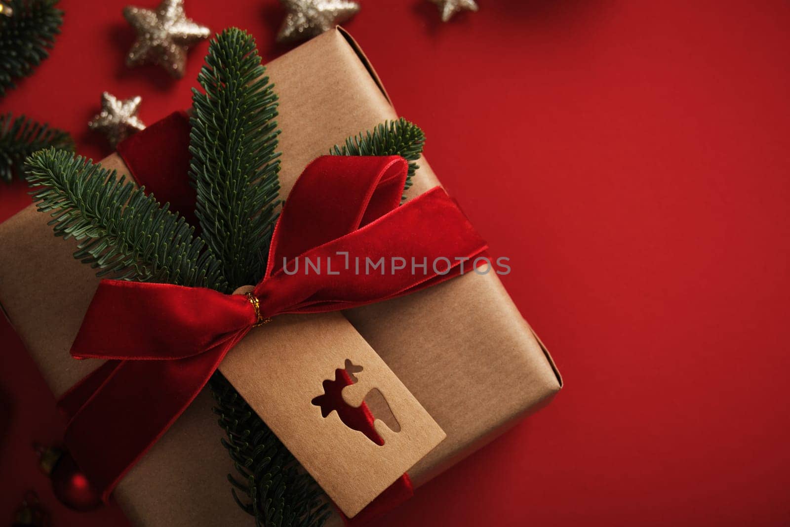 Christmas gift wrapped in kraft paper with red velvet ribbon, a spruce branch and a tag with a silhouette of a reindeer on a red background.