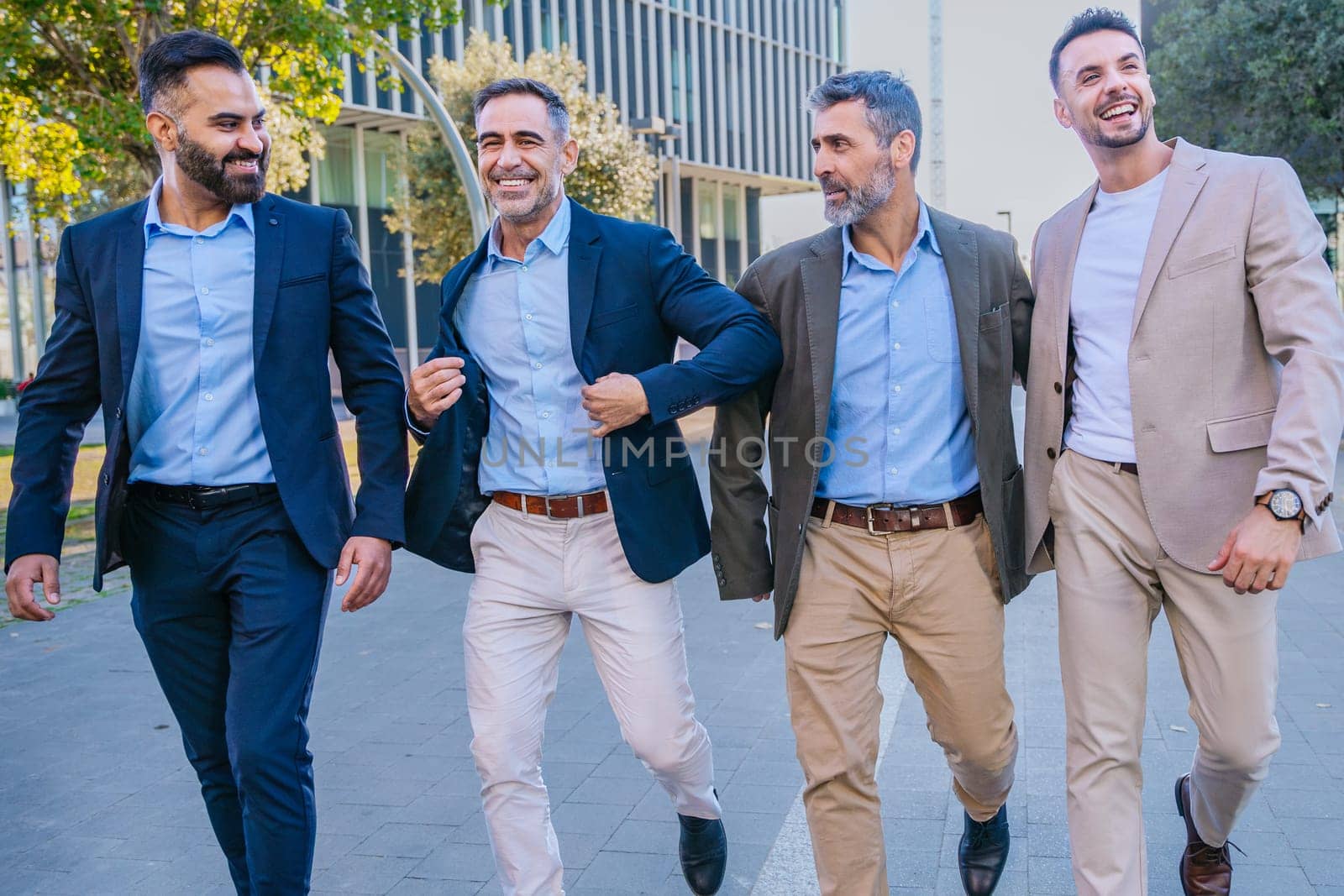 Four businessmen in smart casual and formal attire walking outdoors in an urban setting, smiling and engaging with each other, showcasing teamwork and camaraderie.