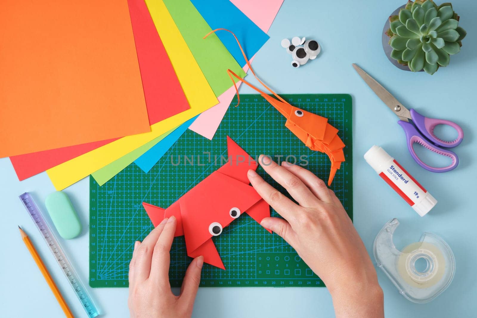 Top view of paper cutting tools, scissors, cutter and paper crafts. Women's hands fixing the final folds for the crab toy. DIY project concept for kids. Flat lay