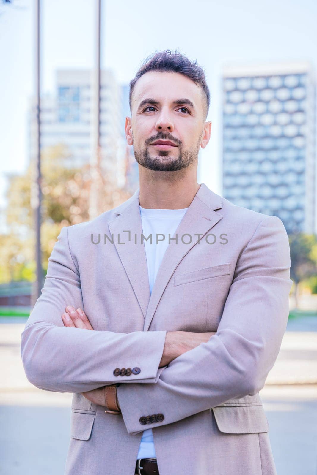 Portrait of a young businessman in a beige blazer standing outdoors in an urban setting with arms crossed, exuding confidence and professionalism