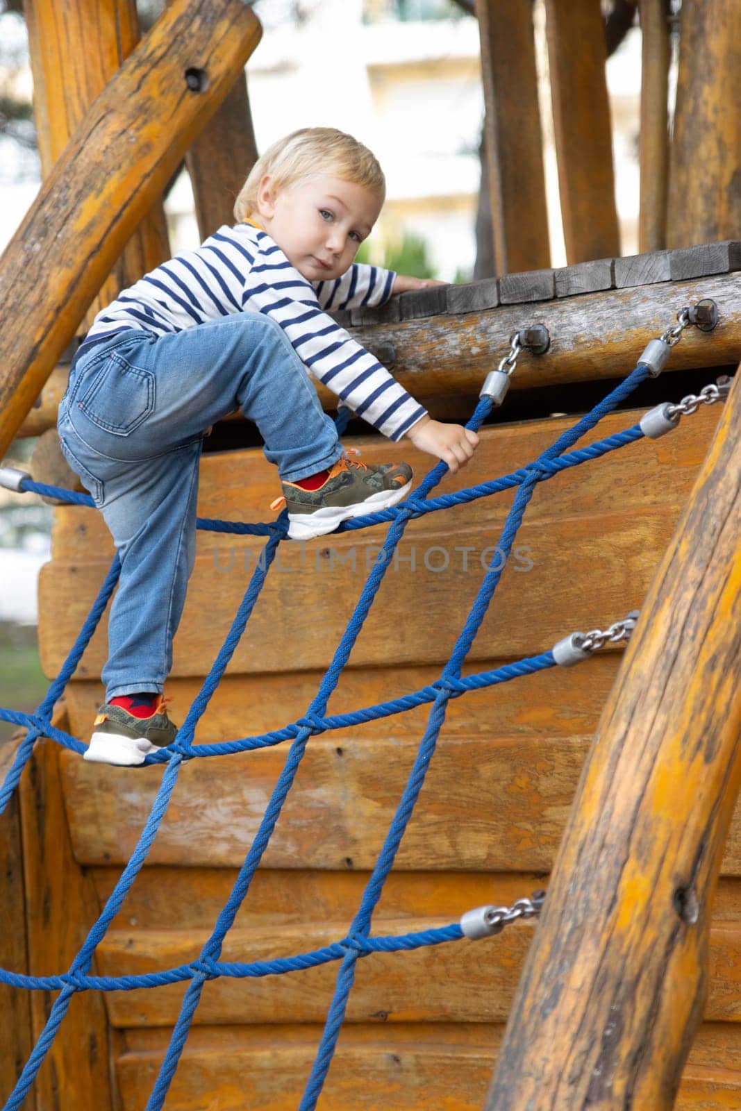 Cute little blonde boy playing on the playground - climbing a rope ladder and looking in the camera. Vertical shot