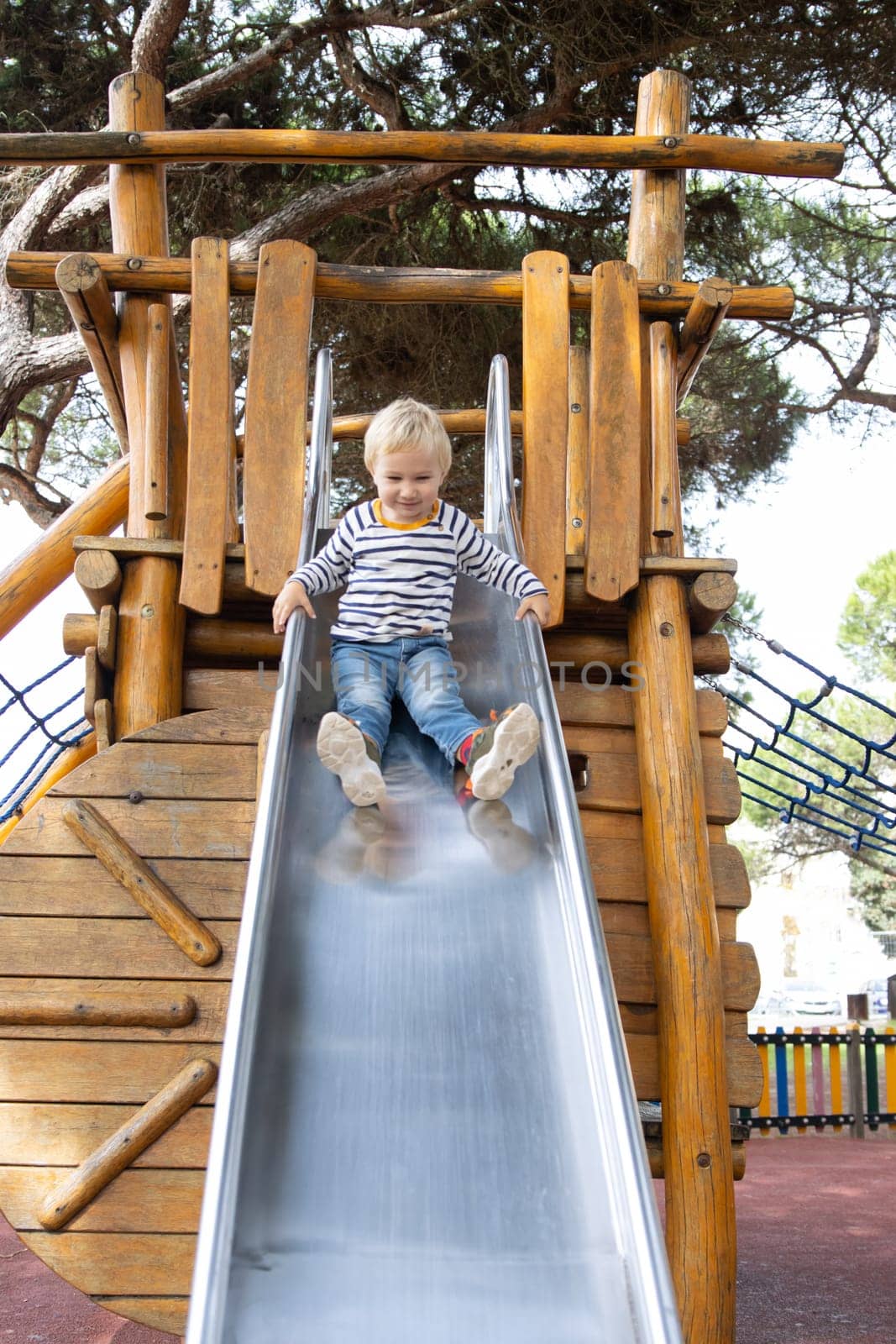 Smiling little blonde boy slide down a slide on the playground. Vertical shot