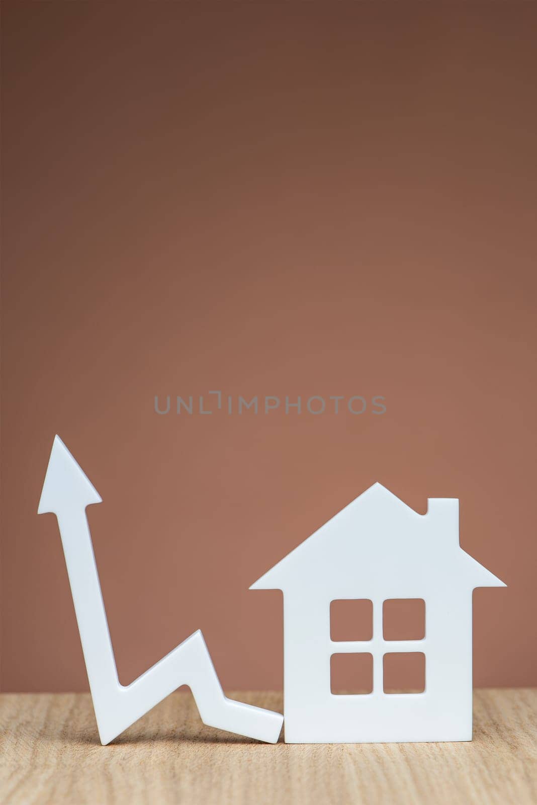 Increasing the cost of renting a home. Banner with a model of a white house and a graphic arrow pointing up close-up on a brown background. Vertical photo with copy space. by SERSOL