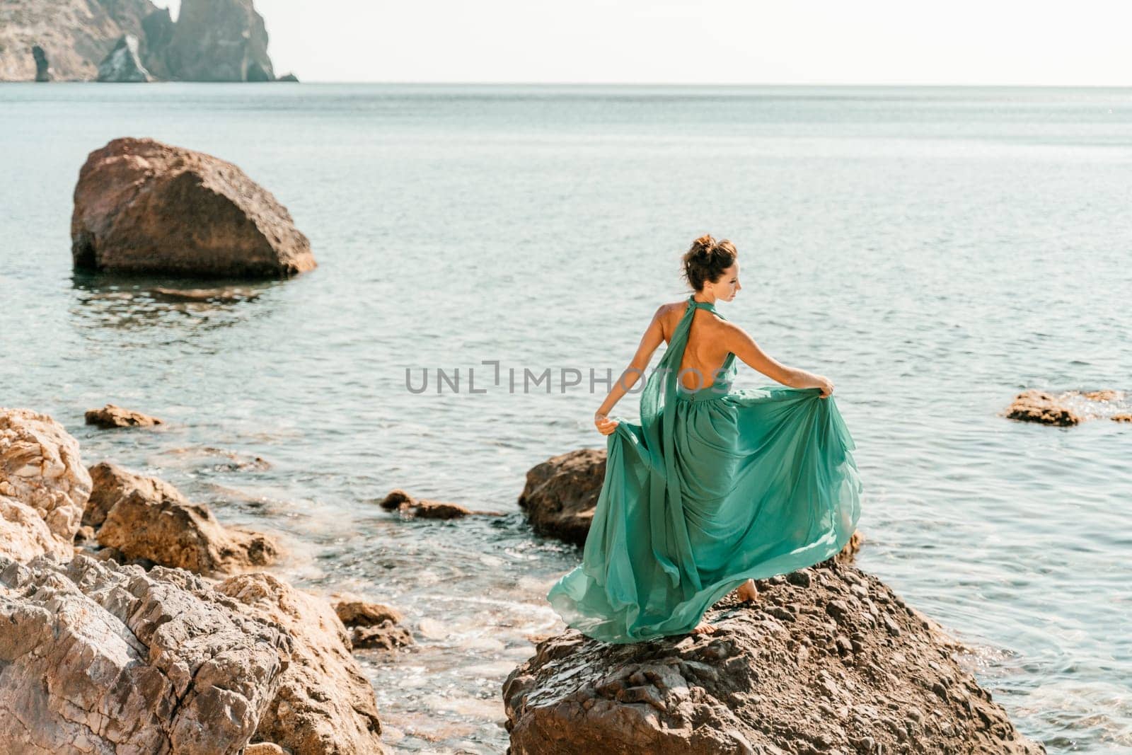 Woman green dress sea. Woman in a long mint dress posing on a beach with rocks on sunny day. Girl on the nature on blue sky background