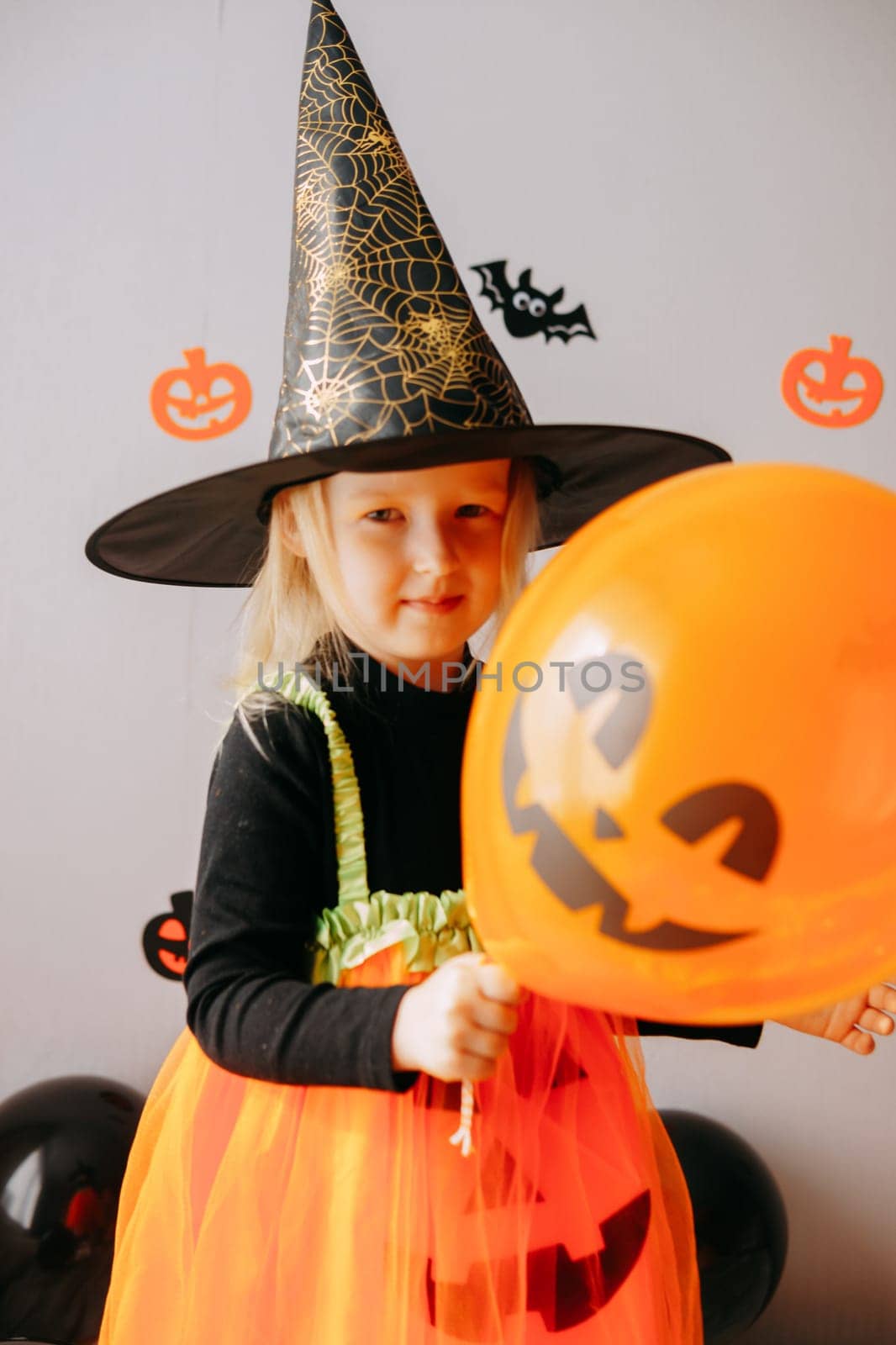 Children's Halloween - a girl in a witch hat and a carnival costume with airy orange and black balloons at home. Ready to celebrate Halloween