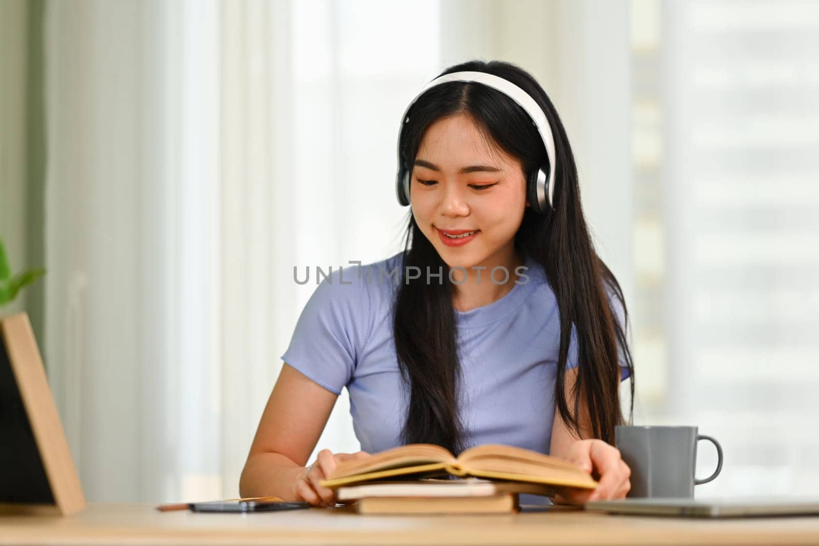 Charming young Asian girl wearing headphone and reading interesting book in bright living room.