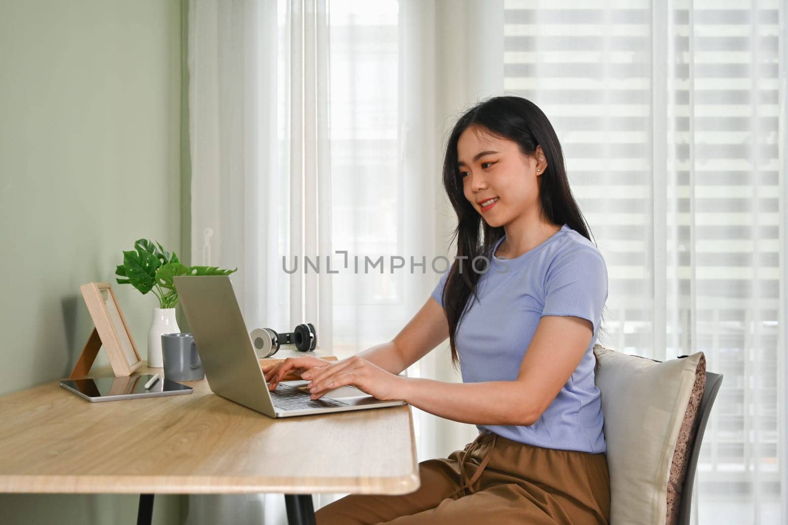 Pleased young asian woman in casual clothes using laptop on wooden desk at home.