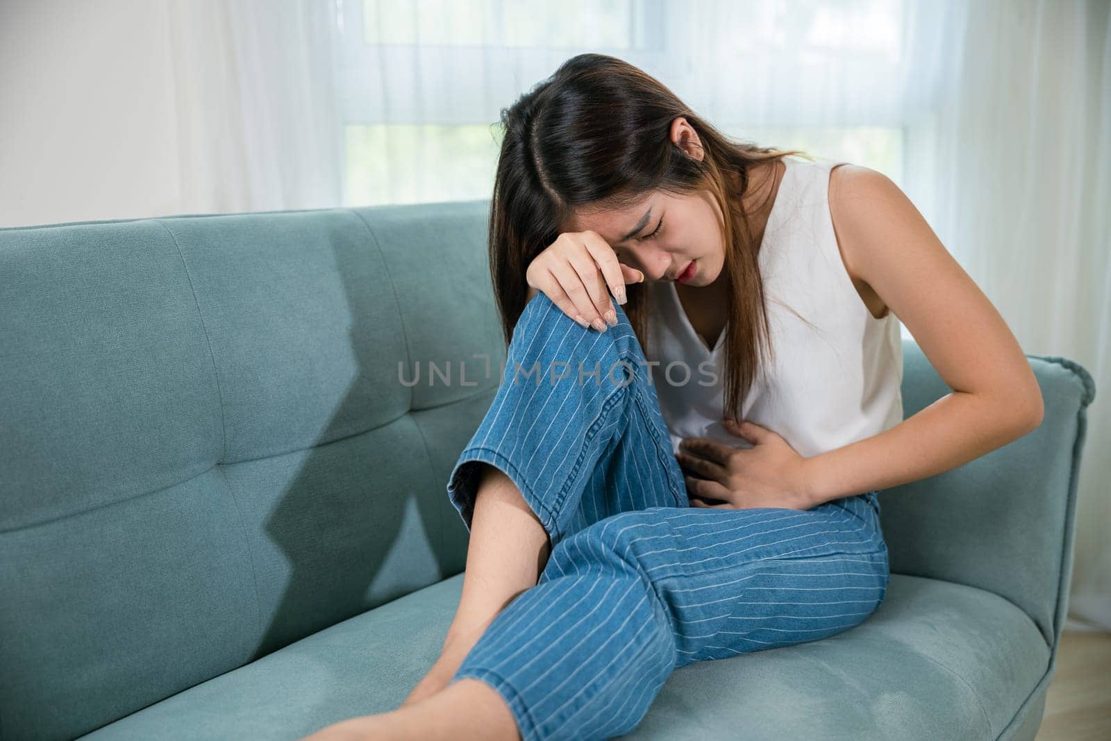 Asian young woman unhappy closing eyes with stomach ache on sofa in living room, female pain lying on couch at home, suffering from strong abdominal painful colon problem feeling unwell