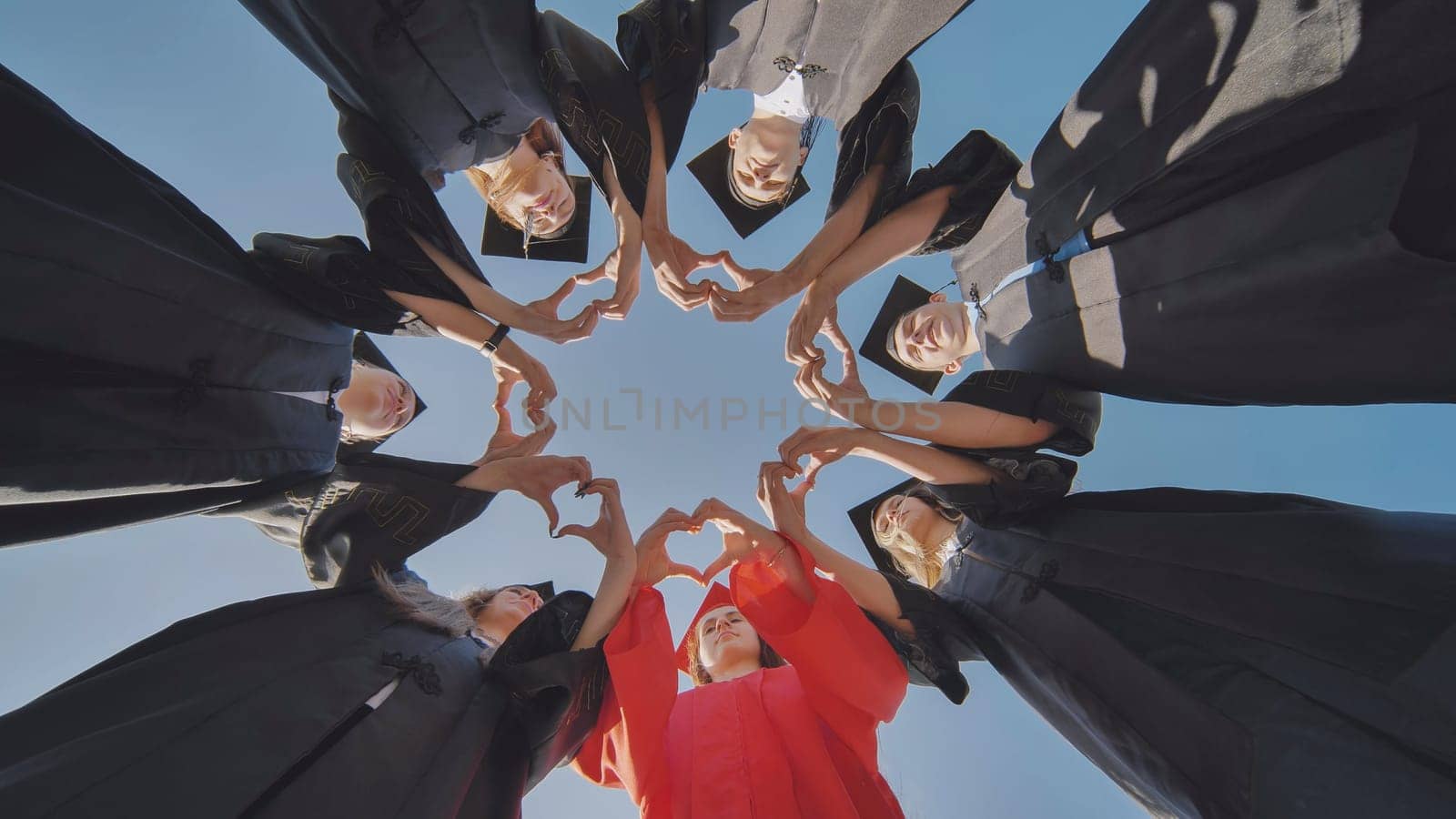 A group of seven college graduates make a heart shape from their hands while standing in a circle wearing robes. by DovidPro