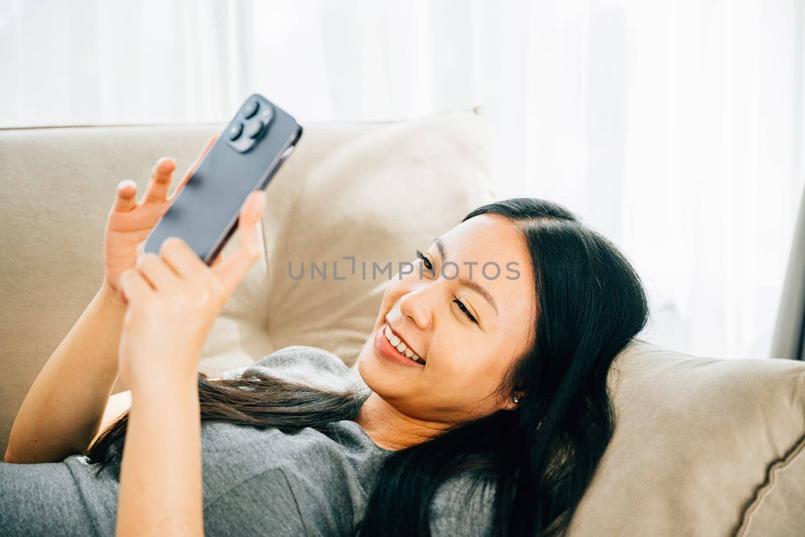 Young Asian woman relaxes on sofa texting on smartphone. Enjoying relaxation chatting and online shopping. Modern technology for communication and connection.