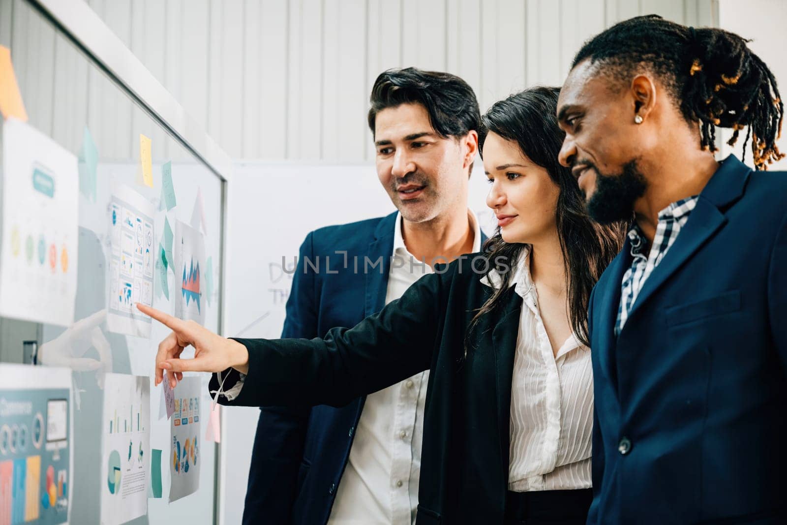 Colleagues in the office come together, utilizing post-it notes on a glass wall for brainstorming. This diverse group embodies teamwork, leadership, and fruitful business discussions.