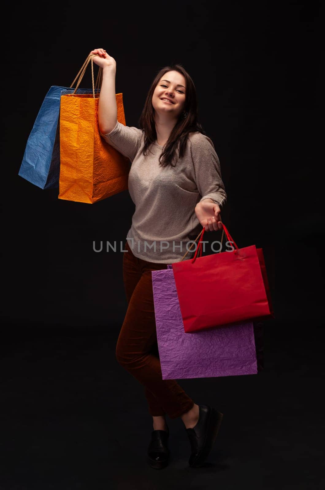 A joyful woman poses with four vibrant shopping bags in a dark studio. She expresses happiness, showcasing the fun of shopping while dressed in casual attire.