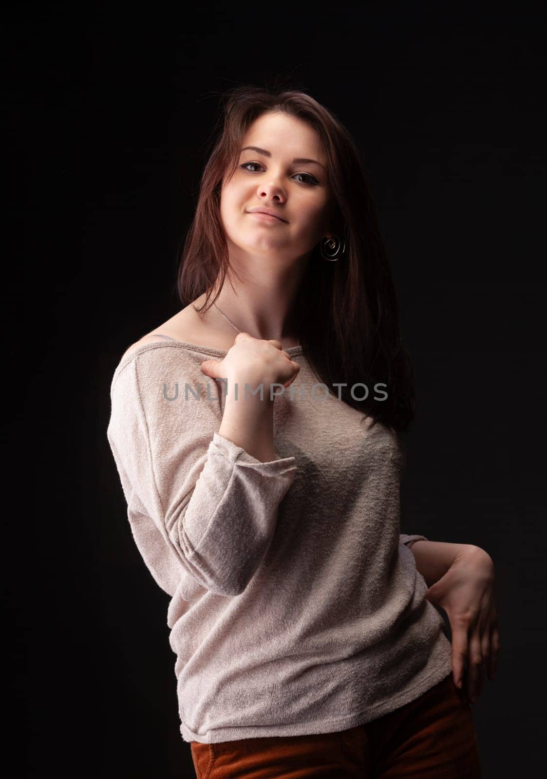 A young woman stands confidently with a playful expression, dressed in a light-colored top. Her stylish pose highlights her personality and fashion sense against a dark backdrop.
