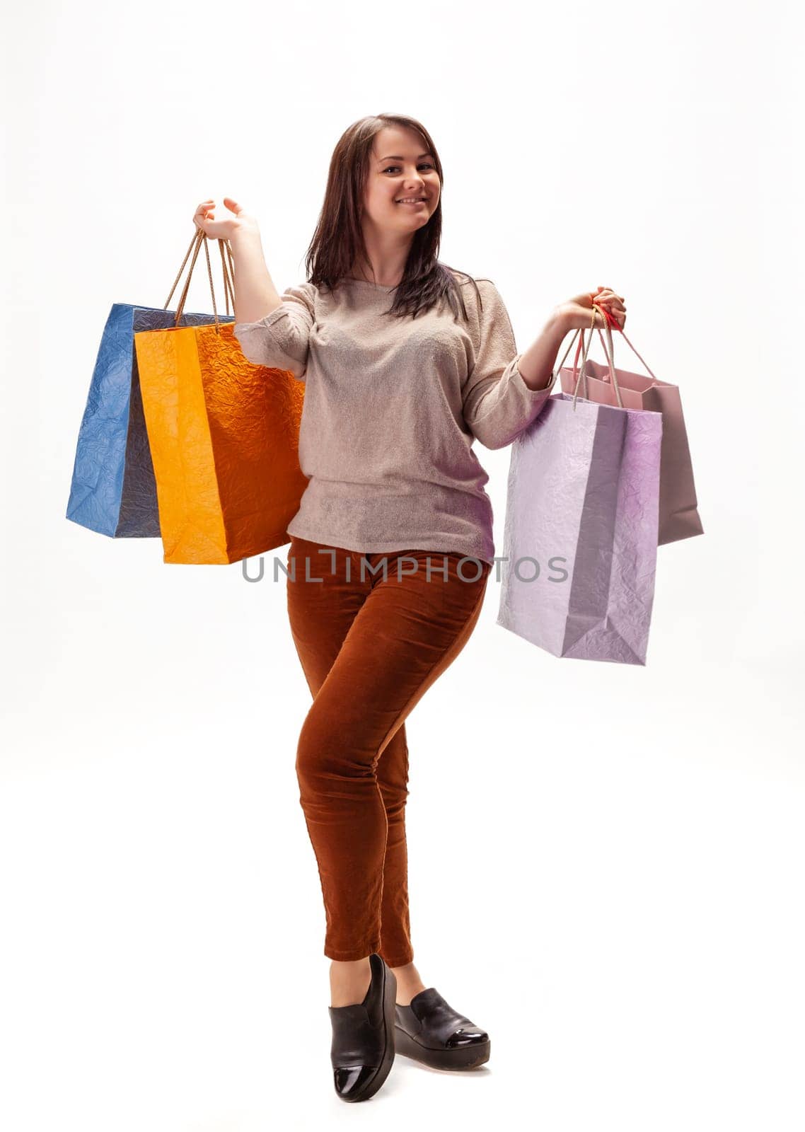 A joyful woman stands confidently in a bright studio, showcasing colorful shopping bags in each hand. She wears a casual outfit, embodying the excitement of shopping.