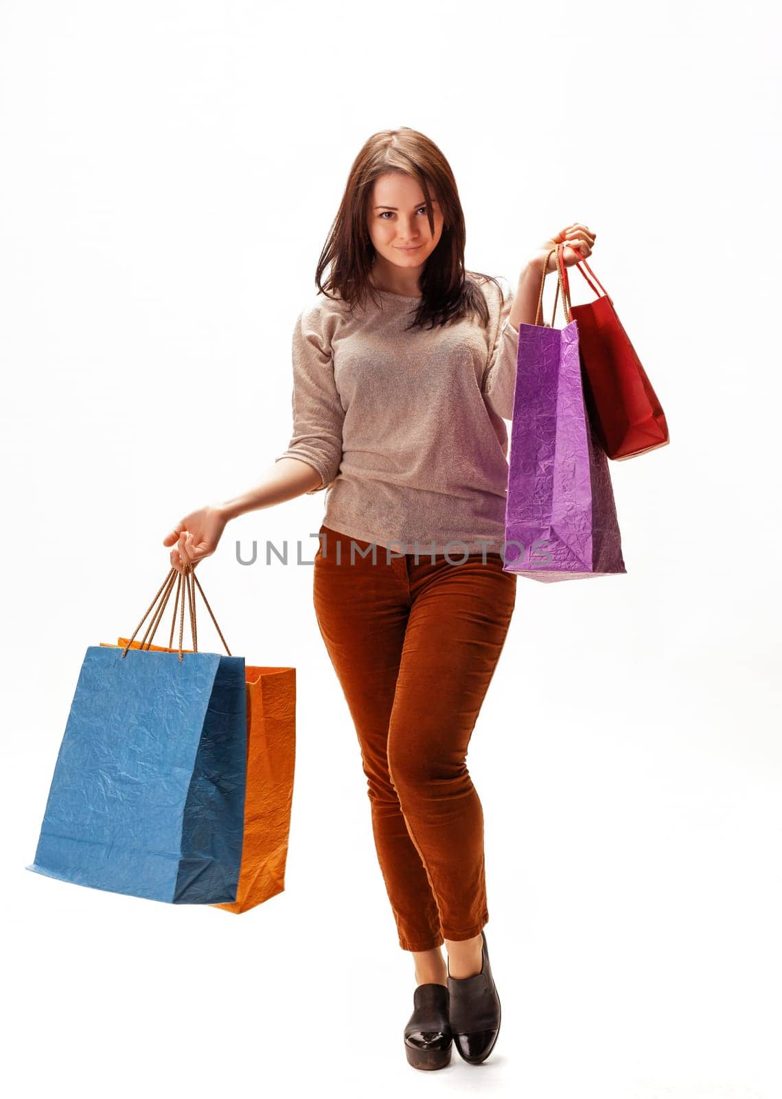 A woman stands confidently with four shopping bags in vibrant colors. She is smiling and dressed casually in a light sweater and brown pants. The bright background enhances her joyful expression.
