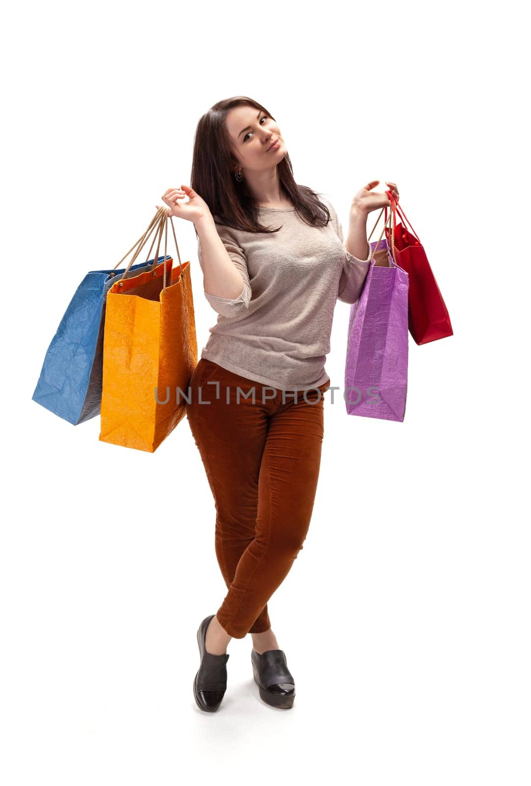 A young woman stands in a bright studio, joyfully holding four colorful shopping bags. She wears a casual outfit and has a playful expression while enjoying her shopping experience.
