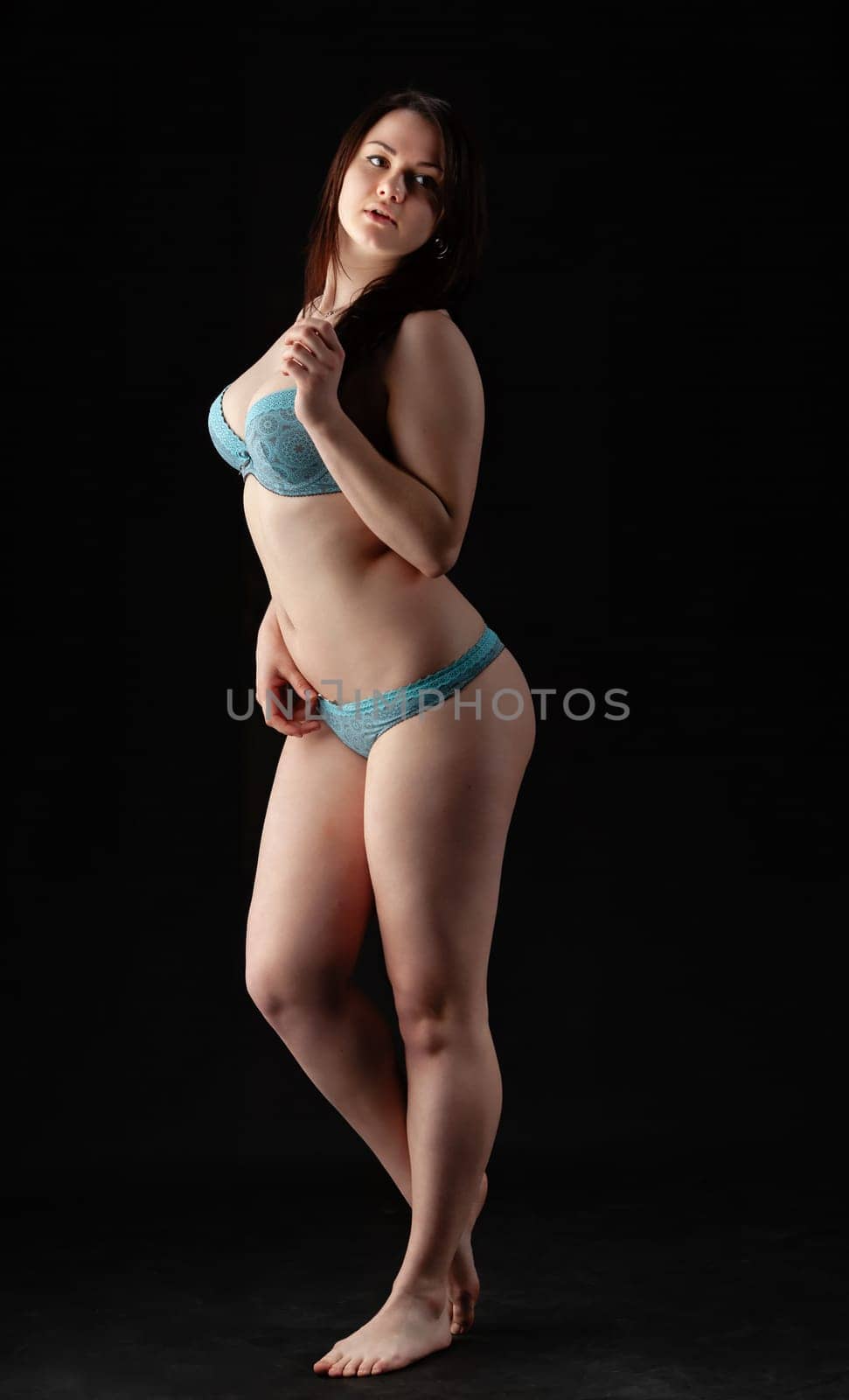 A woman stands elegantly in blue lingerie, showcasing a confident pose. The black background enhances her figure, creating a striking visual contrast during a studio photography session.