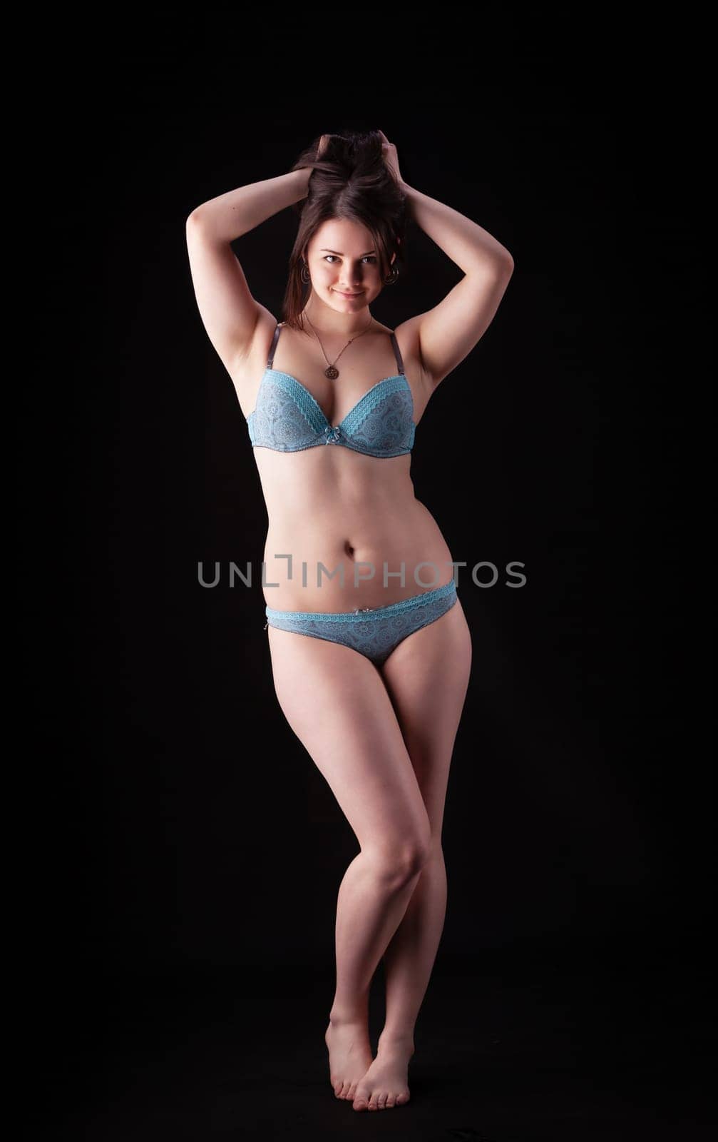 A model stands gracefully with her hands on her head in blue lingerie, showcasing a confident pose and smooth skin.