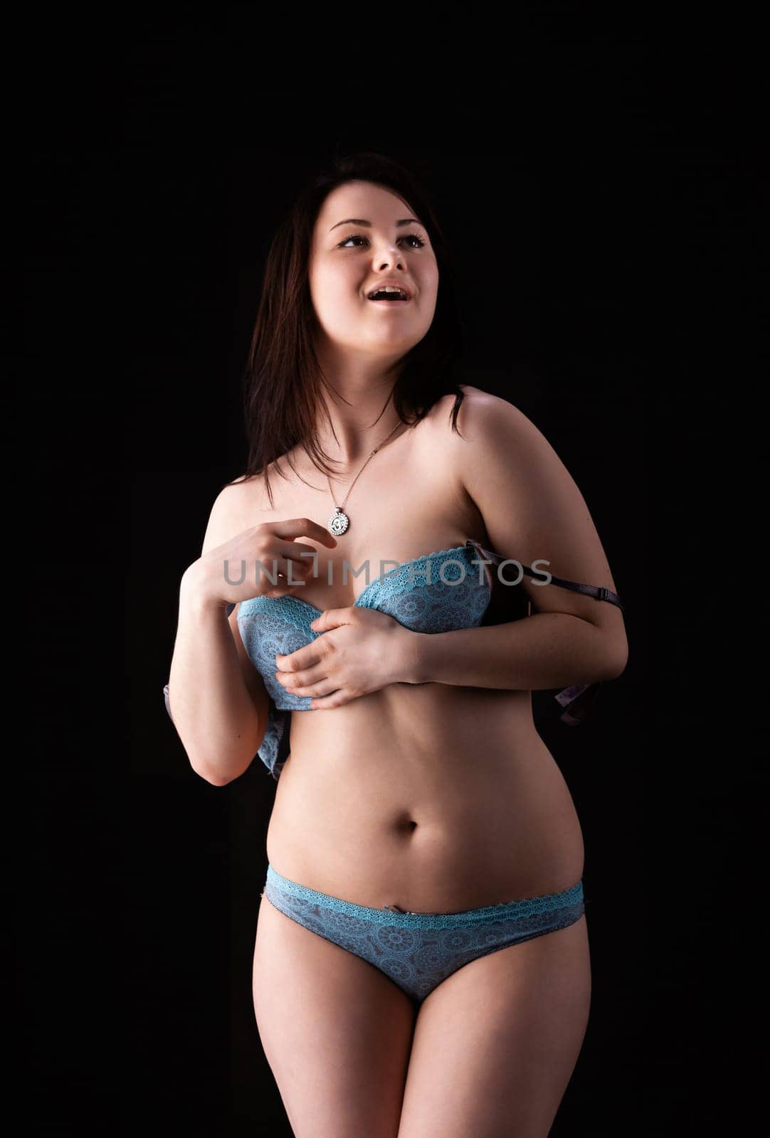 A young woman stands confidently in blue lingerie, smiling and engaging with the camera. The black background emphasizes her playful demeanor.