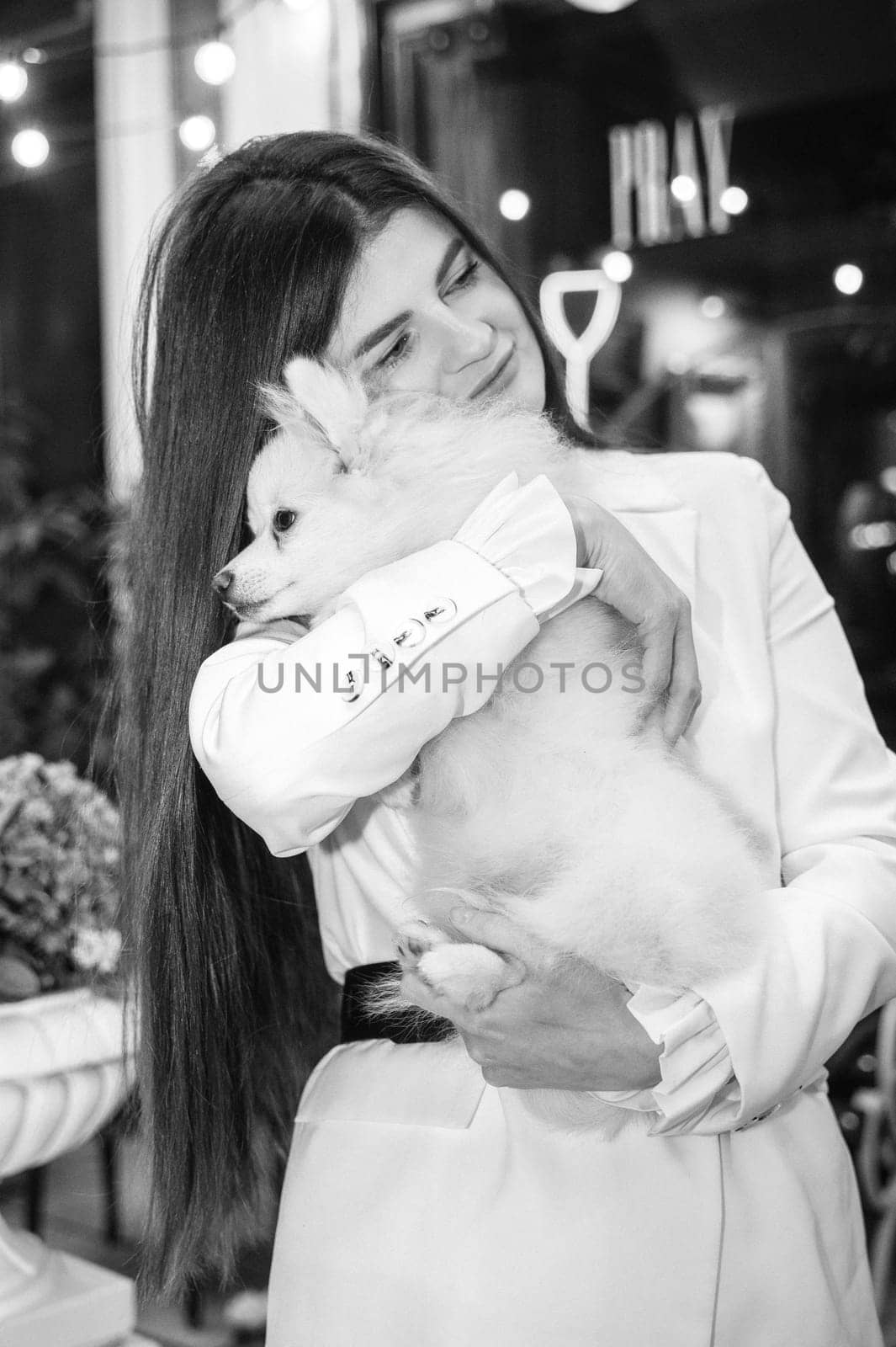A girl holds a white fluffy dog ​​in her arms, Black and white photo. The girl is dressed in a white suit, she has long hair, she loves her dog, a Spitz. The dog is white and fluffy