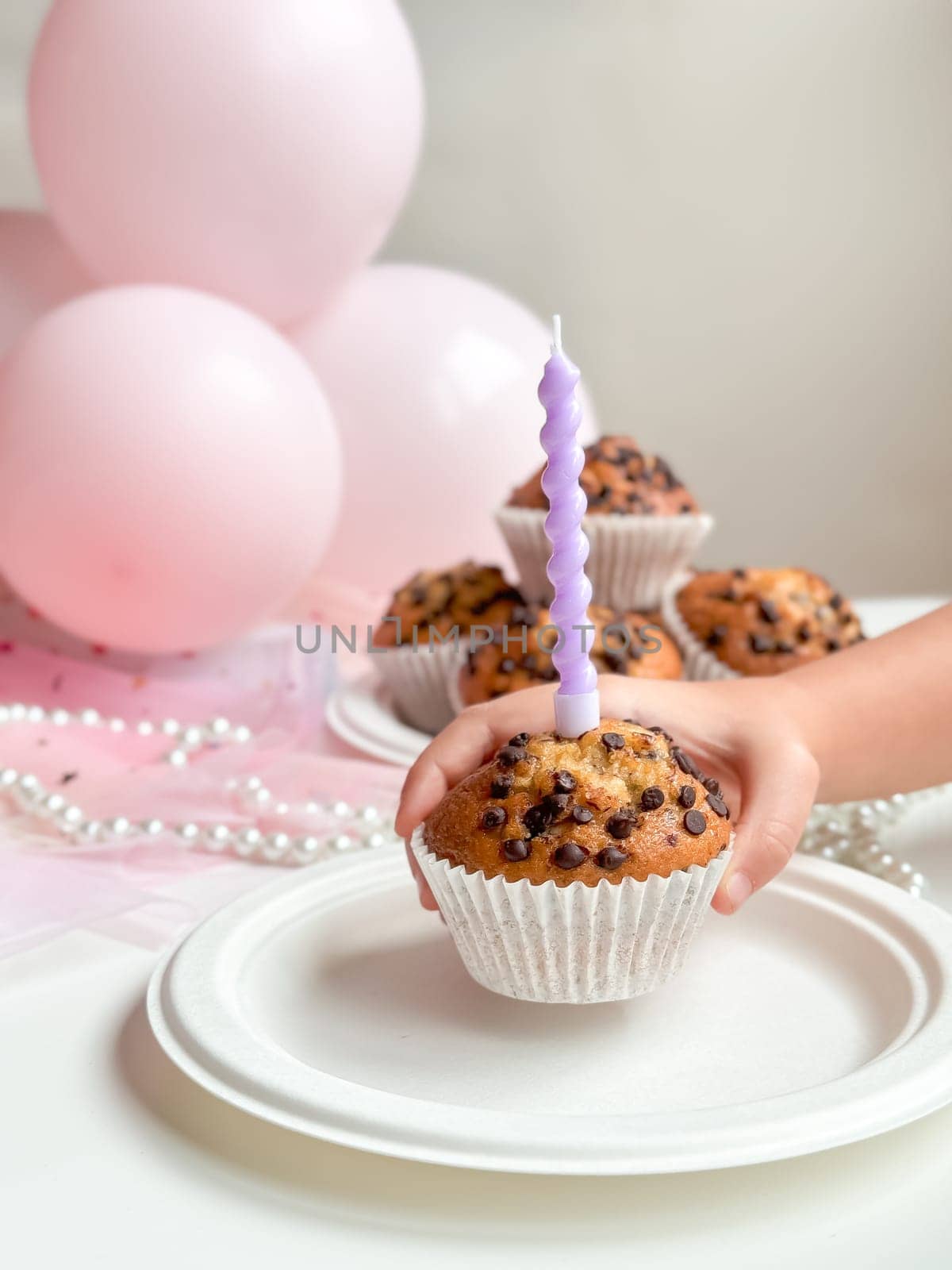 Childs hand holding cupcake with purple candle and chocolate chips next to pink balloons. Birthday concept.