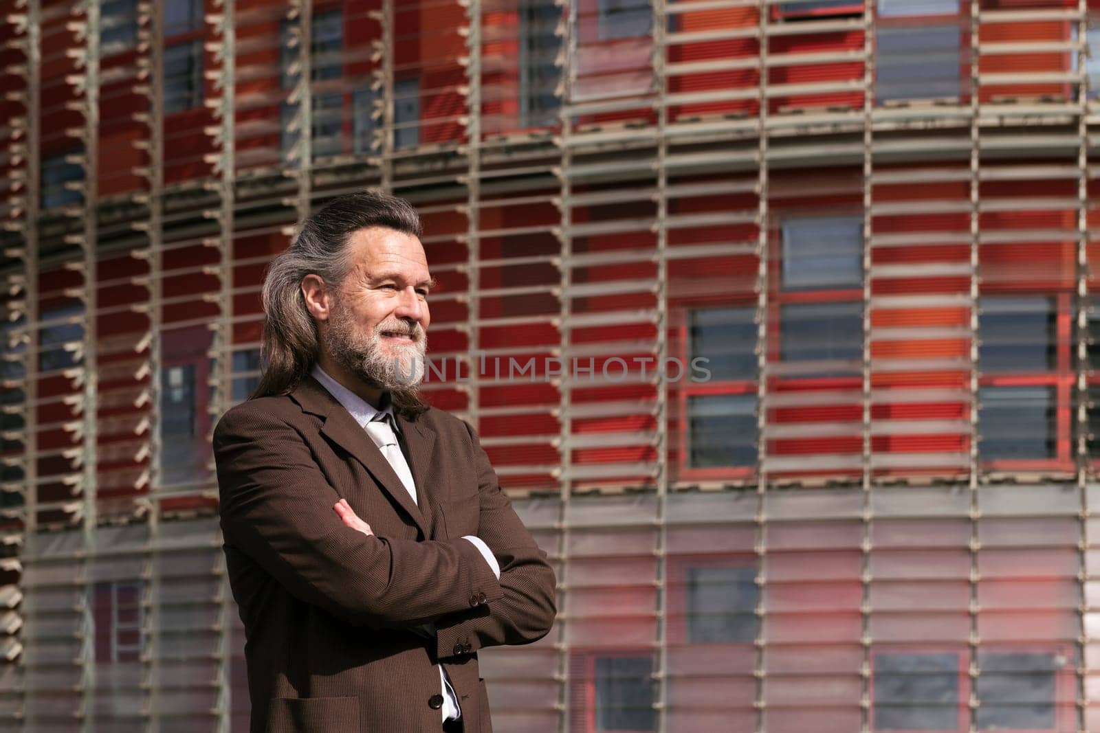 positive middle age male with beard in elegant suit standing with arms crossed and looking away pensive against urban building, concept of business and mature man lifestyle, copyspace for text