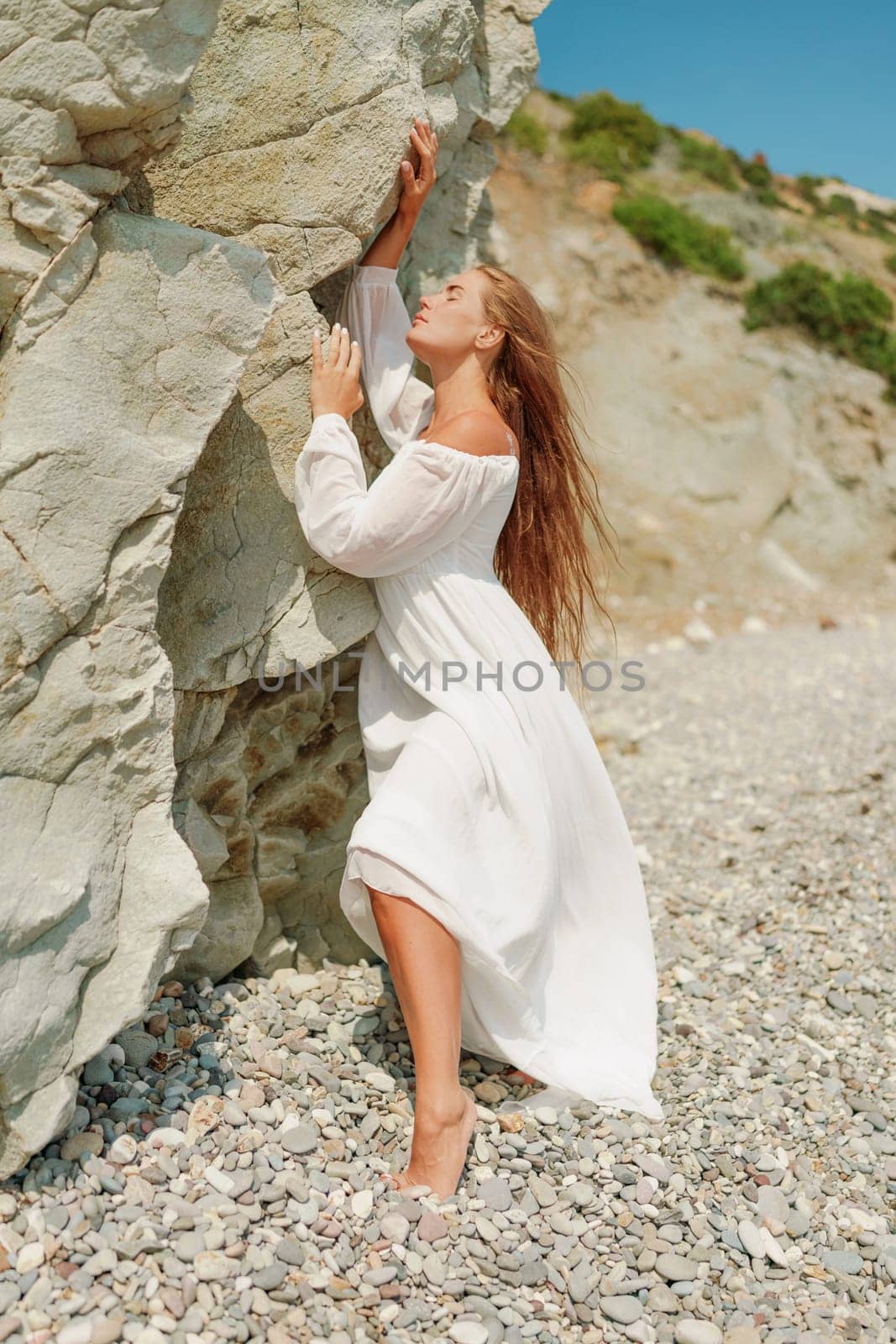 A woman in a white dress is standing on a rocky beach. Concept of serenity and tranquility, as the woman is enjoying the natural beauty of the beach